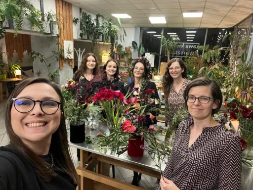 Grupa sześciu uśmiechniętych kobiet pozuje w kwiaciarni pełnej kolorowych kwiatów i roślin doniczkowych.