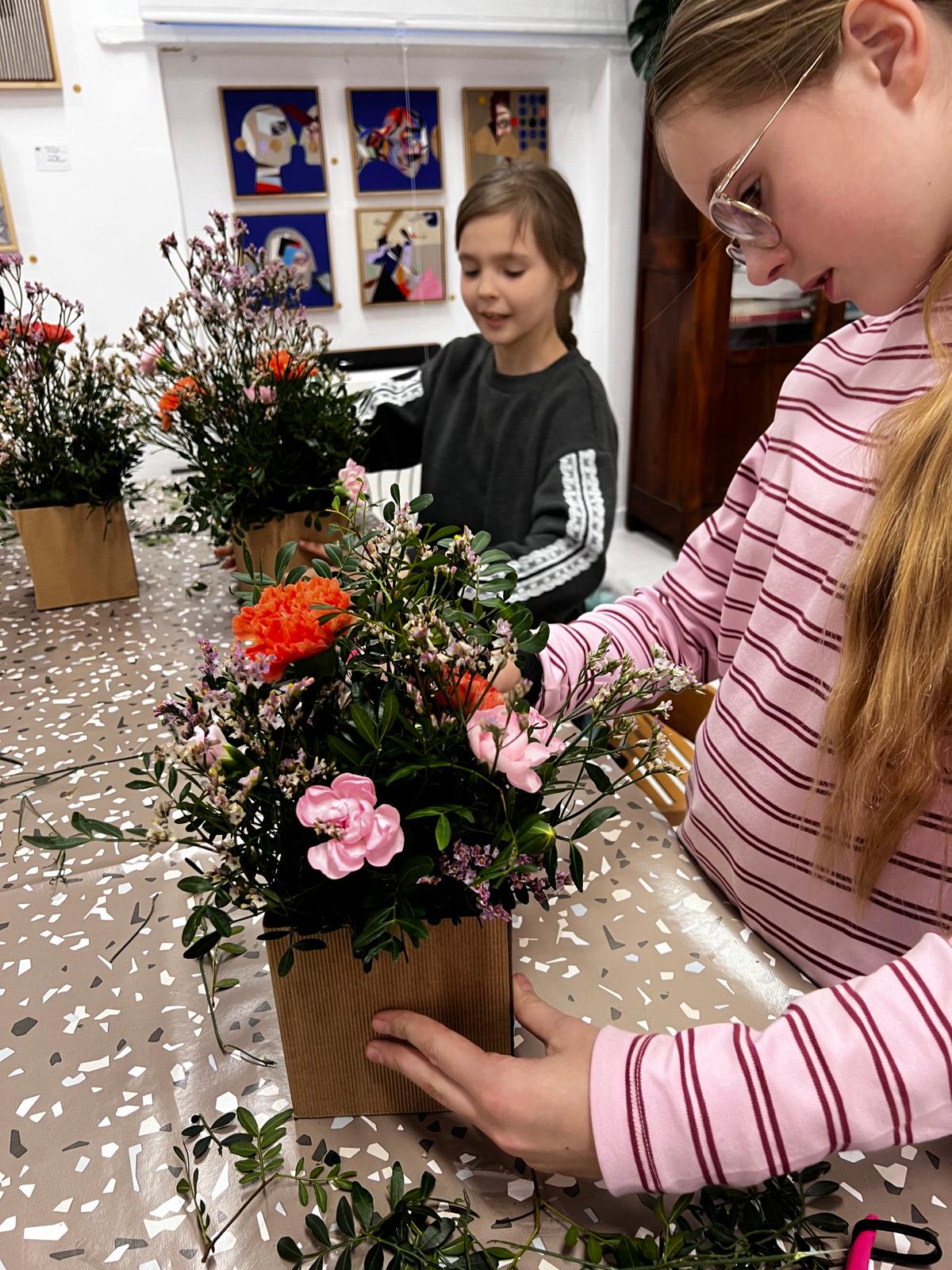 Dwie dziewczyny tworzą kwiatowe kompozycje w papierowych pojemnikach podczas warsztatu florystycznego w galerii sztuki.