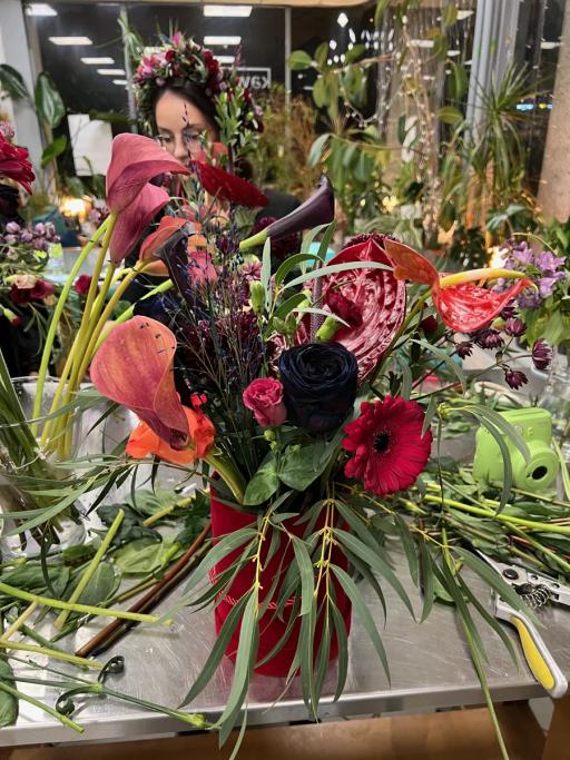 Kwiatowa kompozycja z anthurium, gerberami, różami i liśćmi paproci w odcieniach czerwieni i różu na stole.