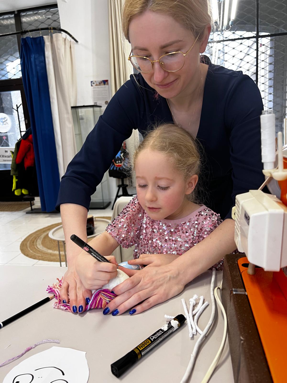 Kobieta w okularach pomaga małej dziewczynie w różowej sukience ozdobić włosy kolorowymi włóknami podczas kreatywnego warsztatu.