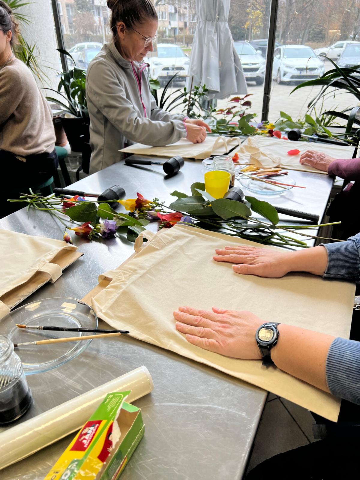 Grupa osób uczestniczy w warsztacie tworzenia bukietów kwiatowych przy stołach z materiałami i narzędziami florystycznymi.