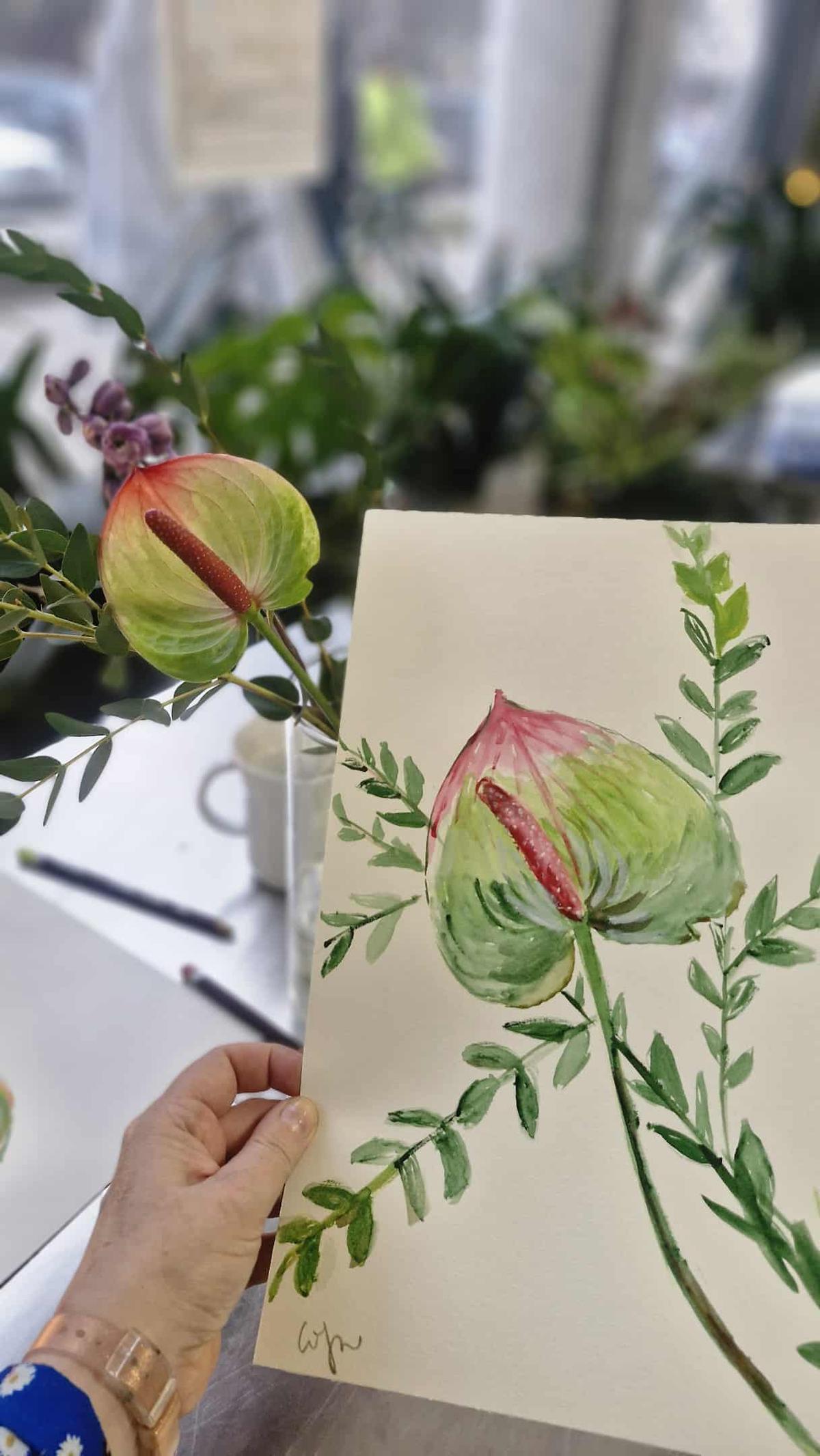 Osoba trzyma akwarelowy rysunek kwiatu z zielonymi liśćmi obok prawdziwej rośliny w doniczce.