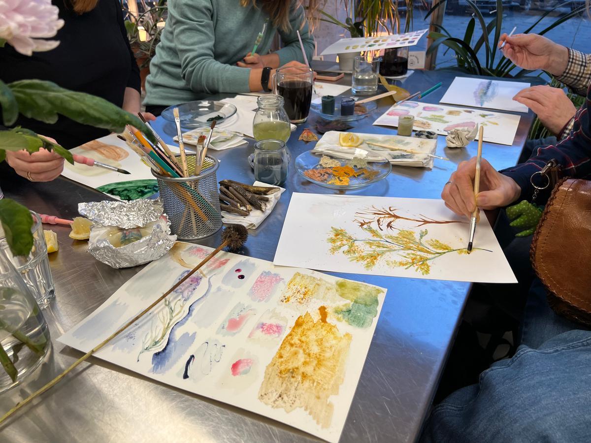 Grupa osób uczestniczy w warsztacie akwareli, malując kwiaty i rośliny przy stole z niezbędnymi materiałami.