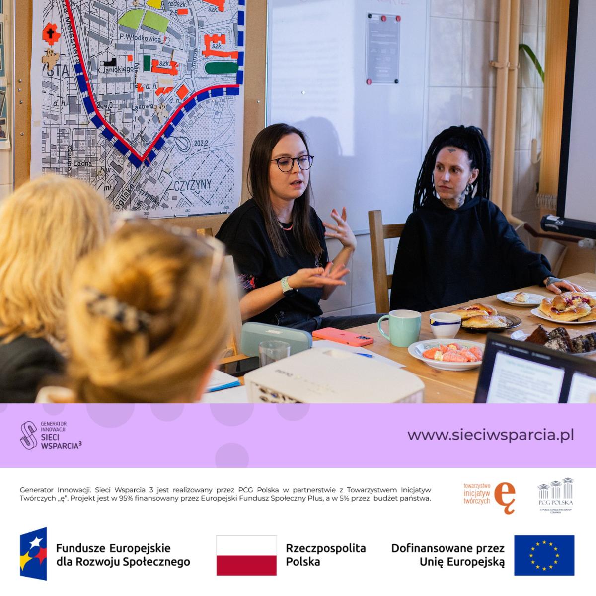 Dwie kobiety dyskutują przy stole podczas spotkania, w tle widoczna mapa miasta z kolorowymi oznaczeniami.