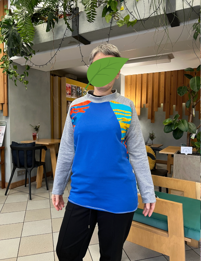 Kobieta w niebieskiej bluzie ze wzorami stoi w kawiarni otoczonej zielonymi roślinami.