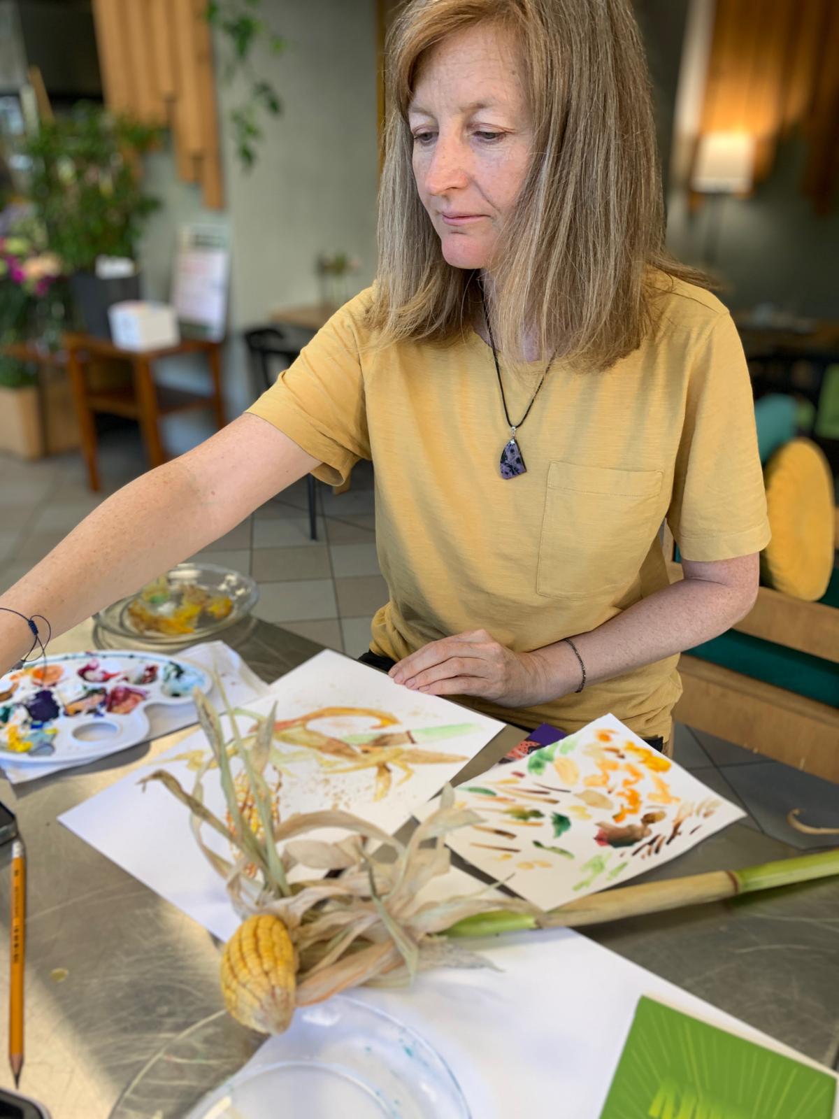 Kobieta w żółtej koszulce przygląda się akwareliowym pinom kwiatów i roślin na stole.