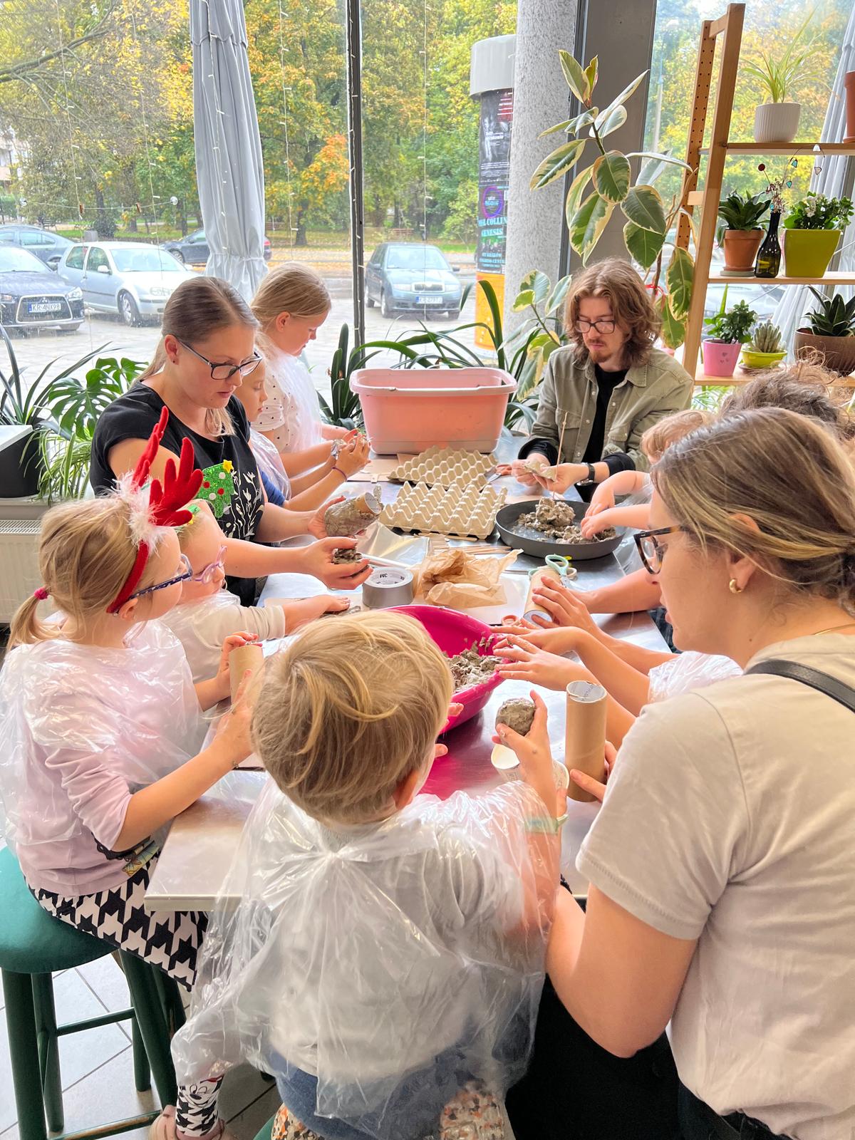 Grupa dzieci uczestniczy w zajęciach plastycznych przy stole, otoczona roślinami doniczkowymi w jasnym pomieszczeniu z dużymi oknami.