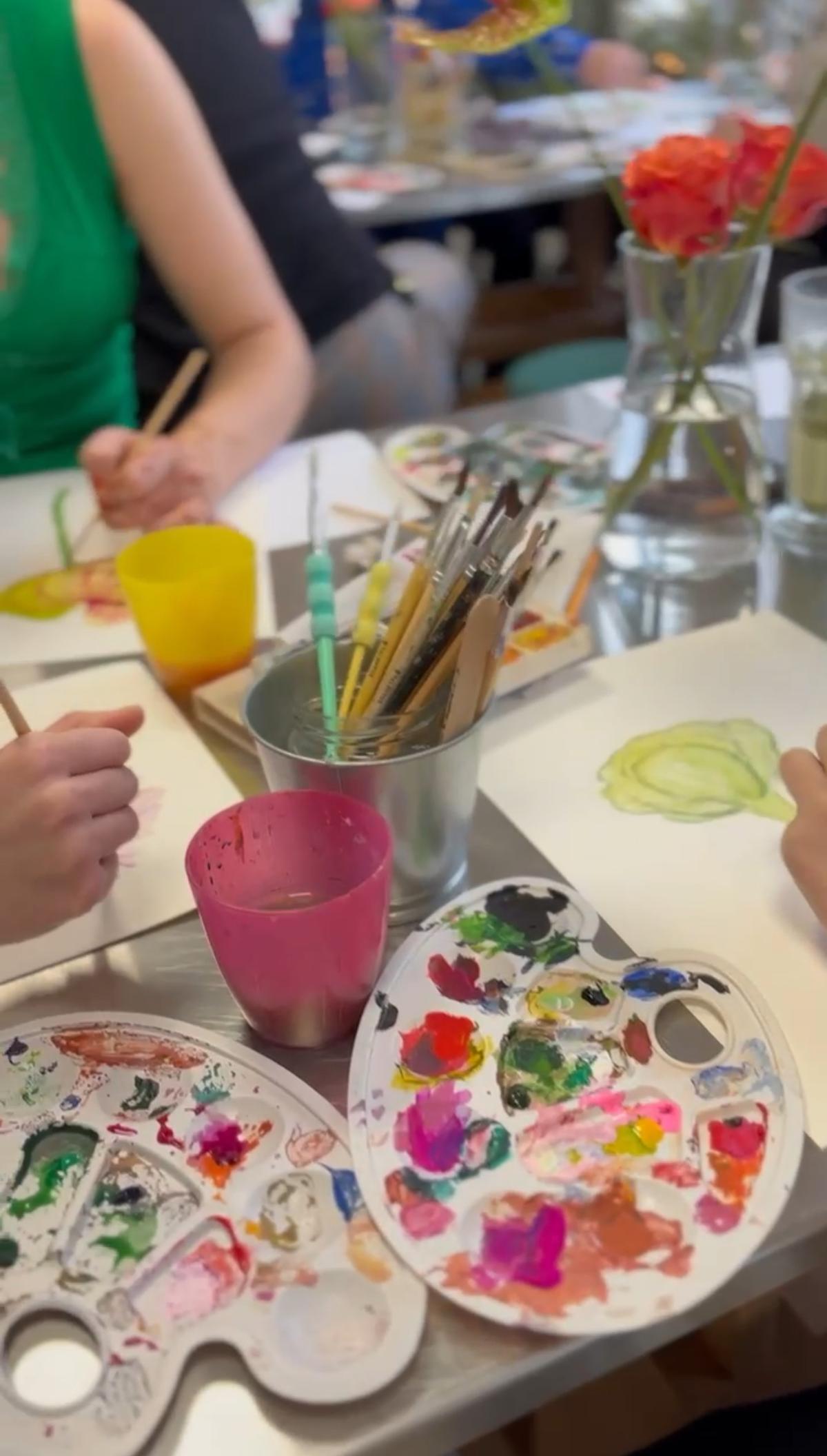 Dzieci uczestniczą w zajęciach plastycznych, malując pędzlami przy stołach z paletami farb i kwiatami w wazonach.