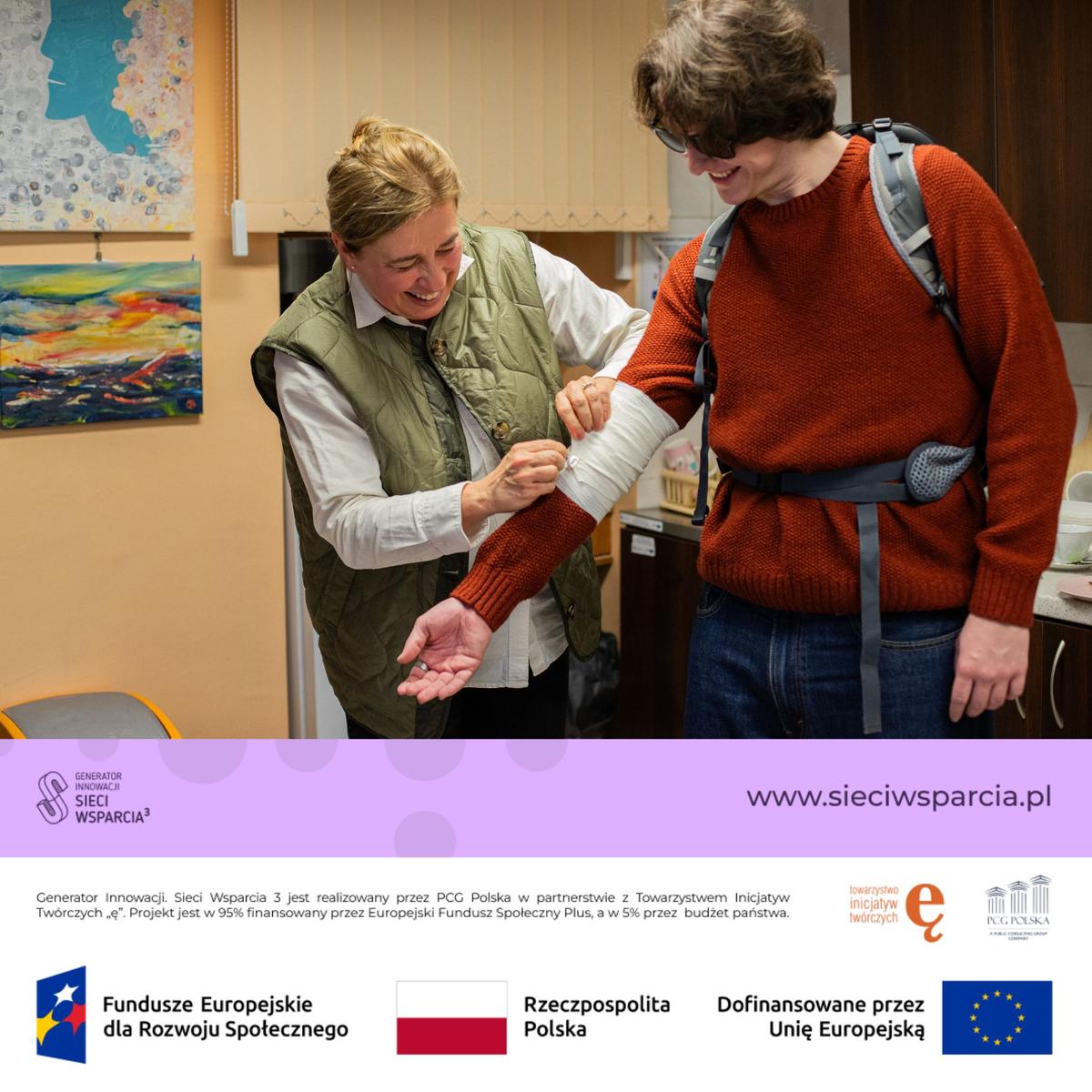 Dwie kobiety w pomieszczeniu omawiają bandaż na ramieniu, przy ścianie z pracami plastycznymi.