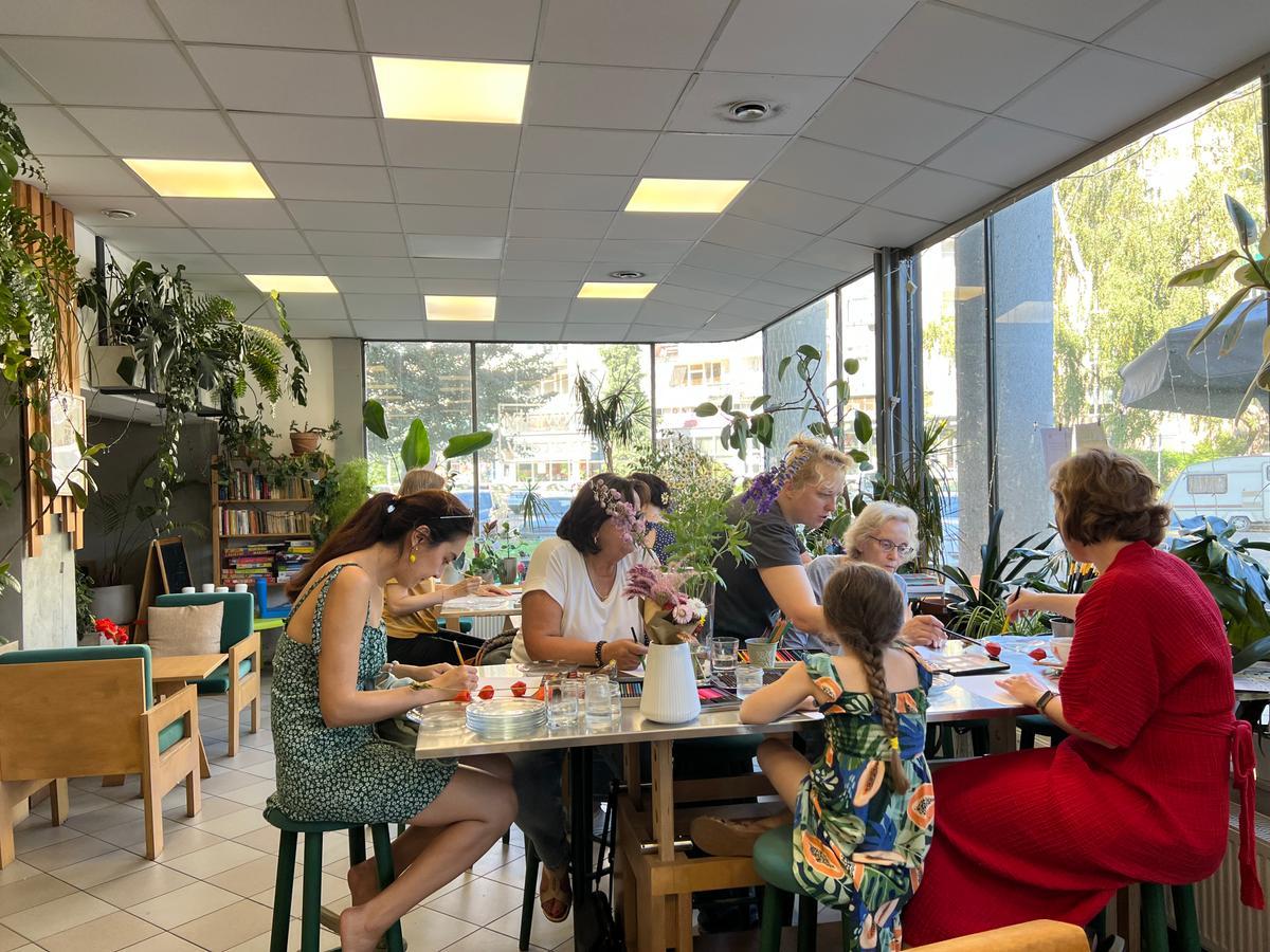 Grupa osób siedzi przy stołach w jasnej sali pełnej roślin, spożywając posiłek w przyjaznym otoczeniu.