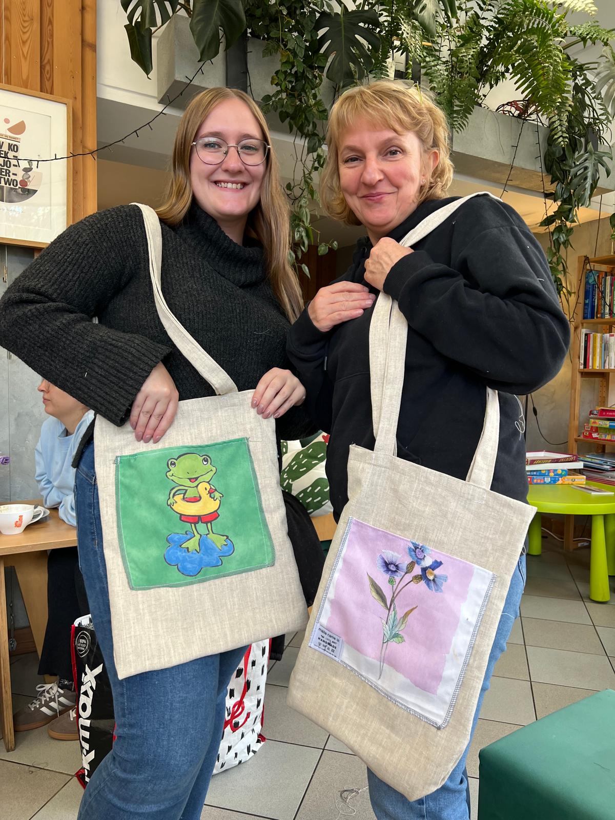 Dwie osoby prezentują torby ze składanymi kieszonkami ozdabianymi kolorowym rysunkiem żaby i kwiatów.