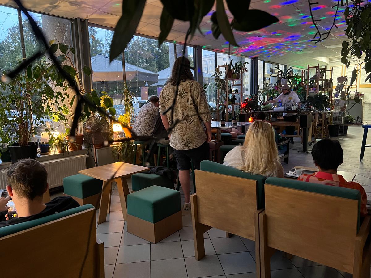 Nowoczesne wnętrze kawiarni lub biura z roślinami, kolorowymi światłami na suficie i kilkoma osobami siedzącymi na drewnianych fotelach.