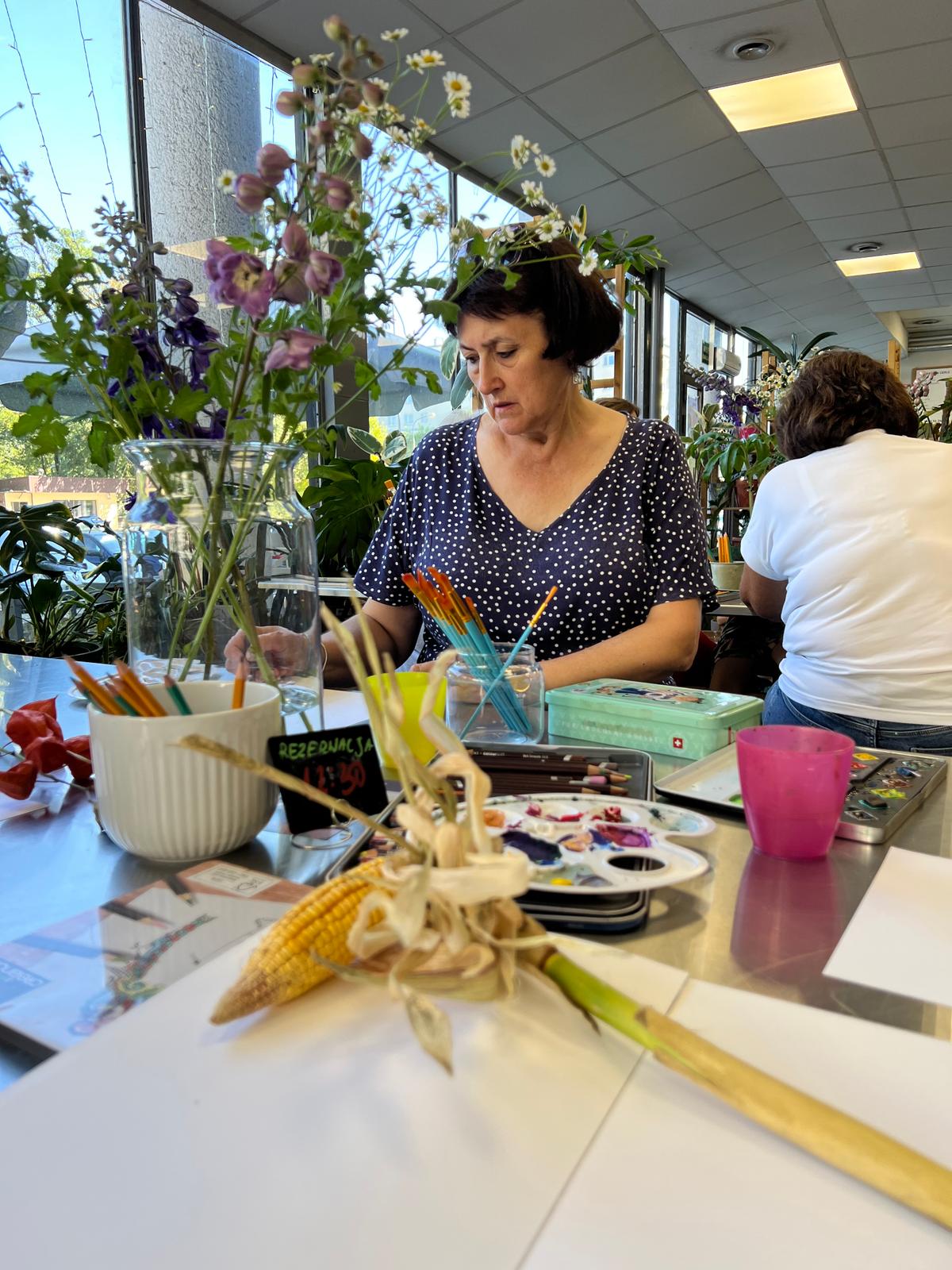 Kobieta w kropkowanej bluzce maluje przy stole pełnym kwiatów, pędzli i materiałów artystycznych w jasnym pomieszczeniu.