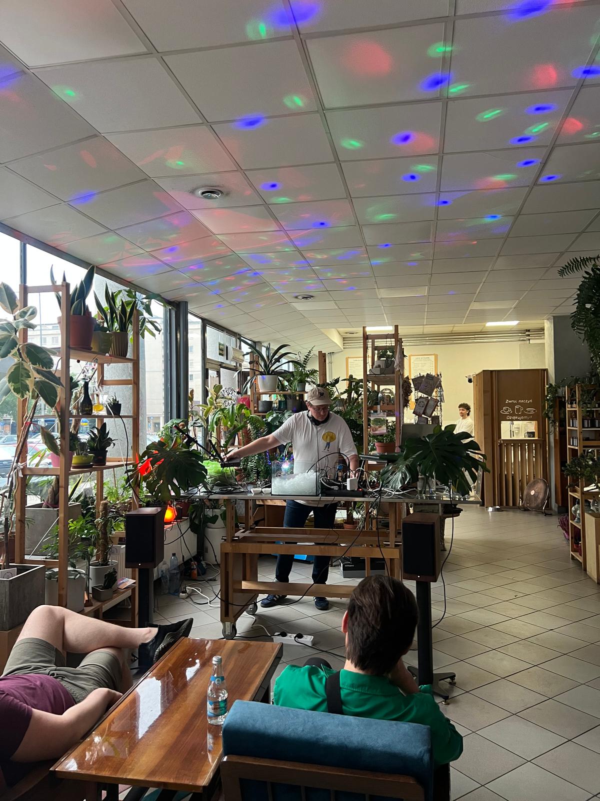 Mężczyzna DJ-uje w zielonej kawiarni pełnej roślin, przy kolorowych lampach sufitowych, podczas gdy goście słuchają muzyki.
