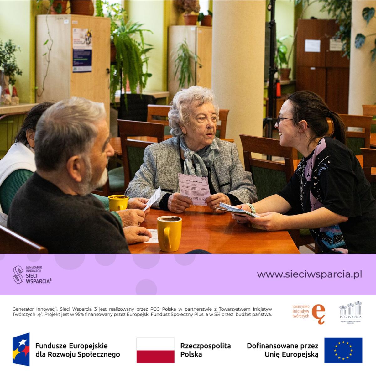 Trzech uczestników rozmawia przy stole w sali spotkań, a starsze osoby trzymają dokumenty projektu.