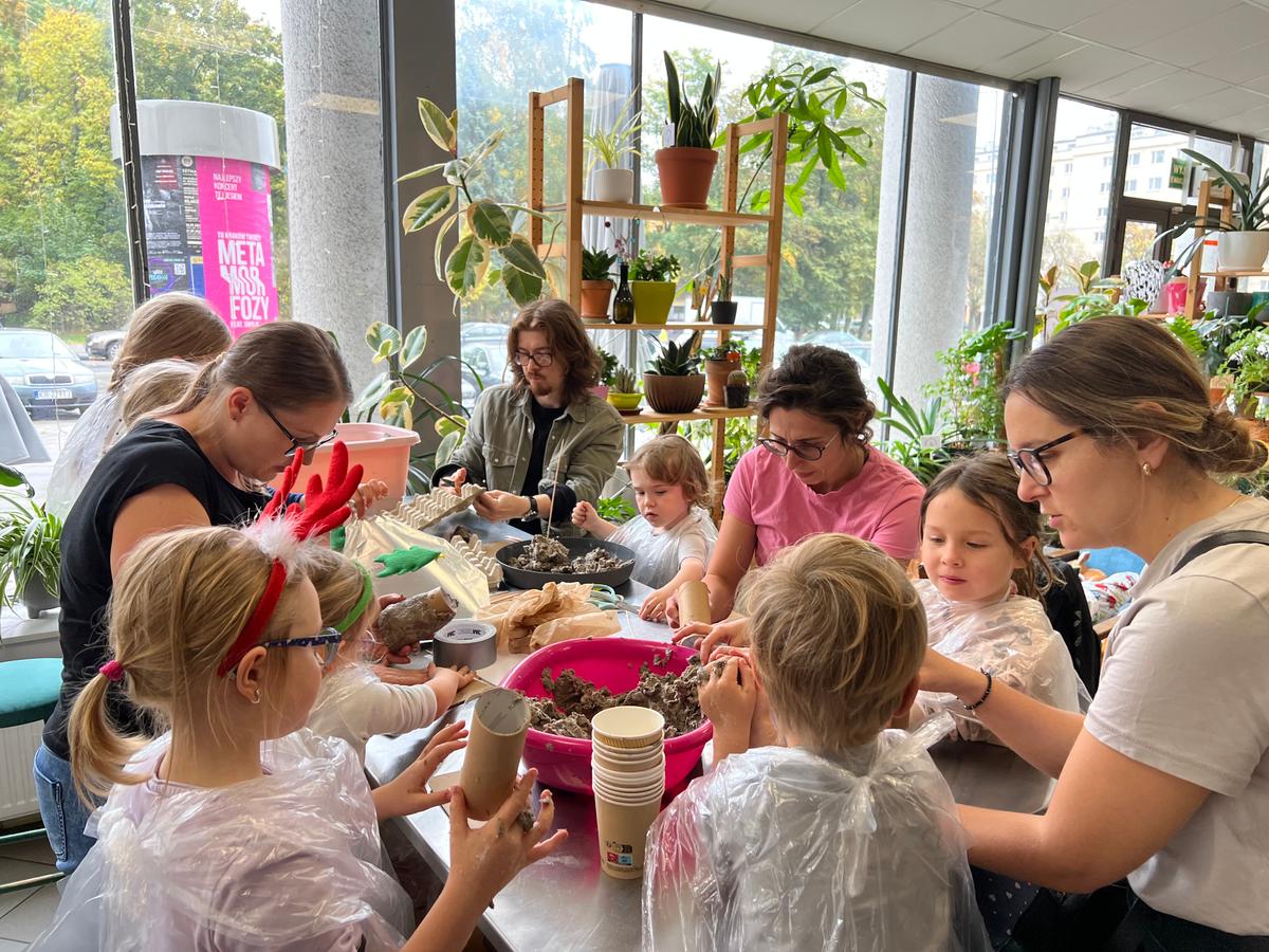 Grupa dzieci i dorosłych uczestniczy w warsztacie sadzenia roślin w nowoczesnym pomieszczeniu pełnym zieleni.