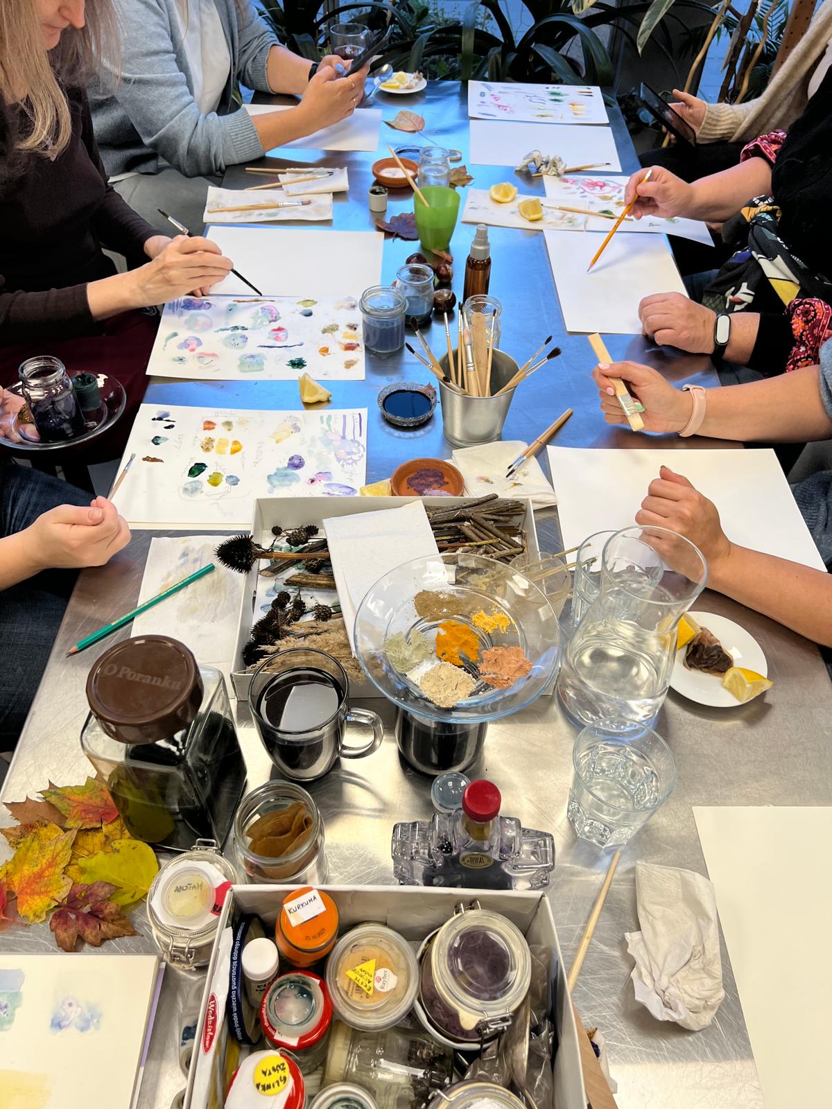 Grupa ludzi siedzi przy stole i uczestniczy w warsztatach malarskich z naturalnymi materiałami.