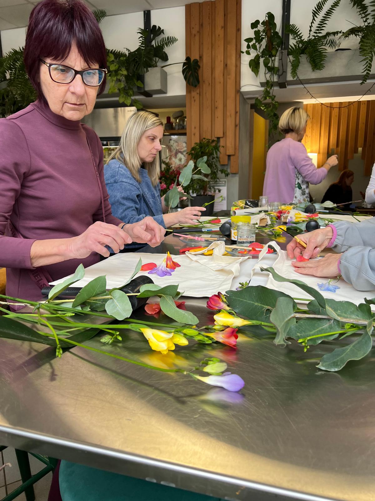 Grupa osób przy stole wykonuje pracę manualną z kwiatami i liśćmi w otoczeniu roślin doniczkowych.