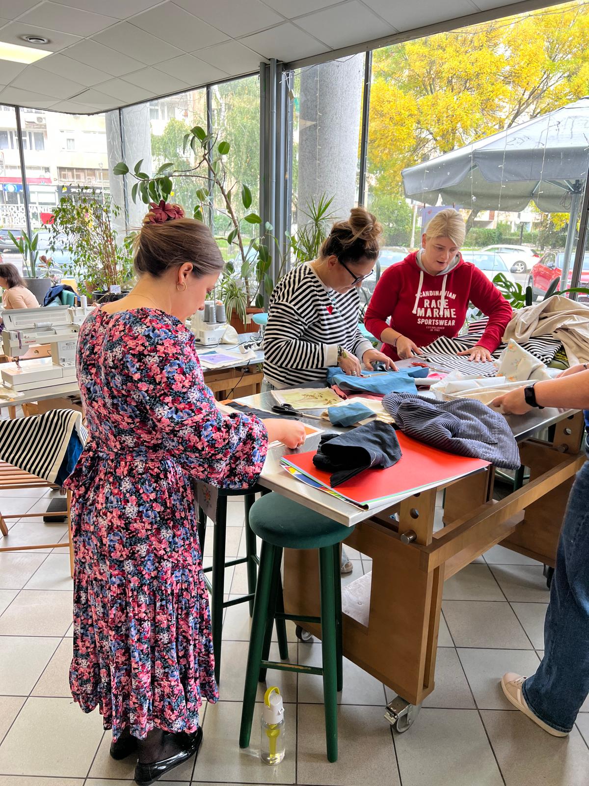 Grupa osób pracuje przy stołach w jasnym, przeszklonym pomieszczeniu, sortując i układając ubrania.