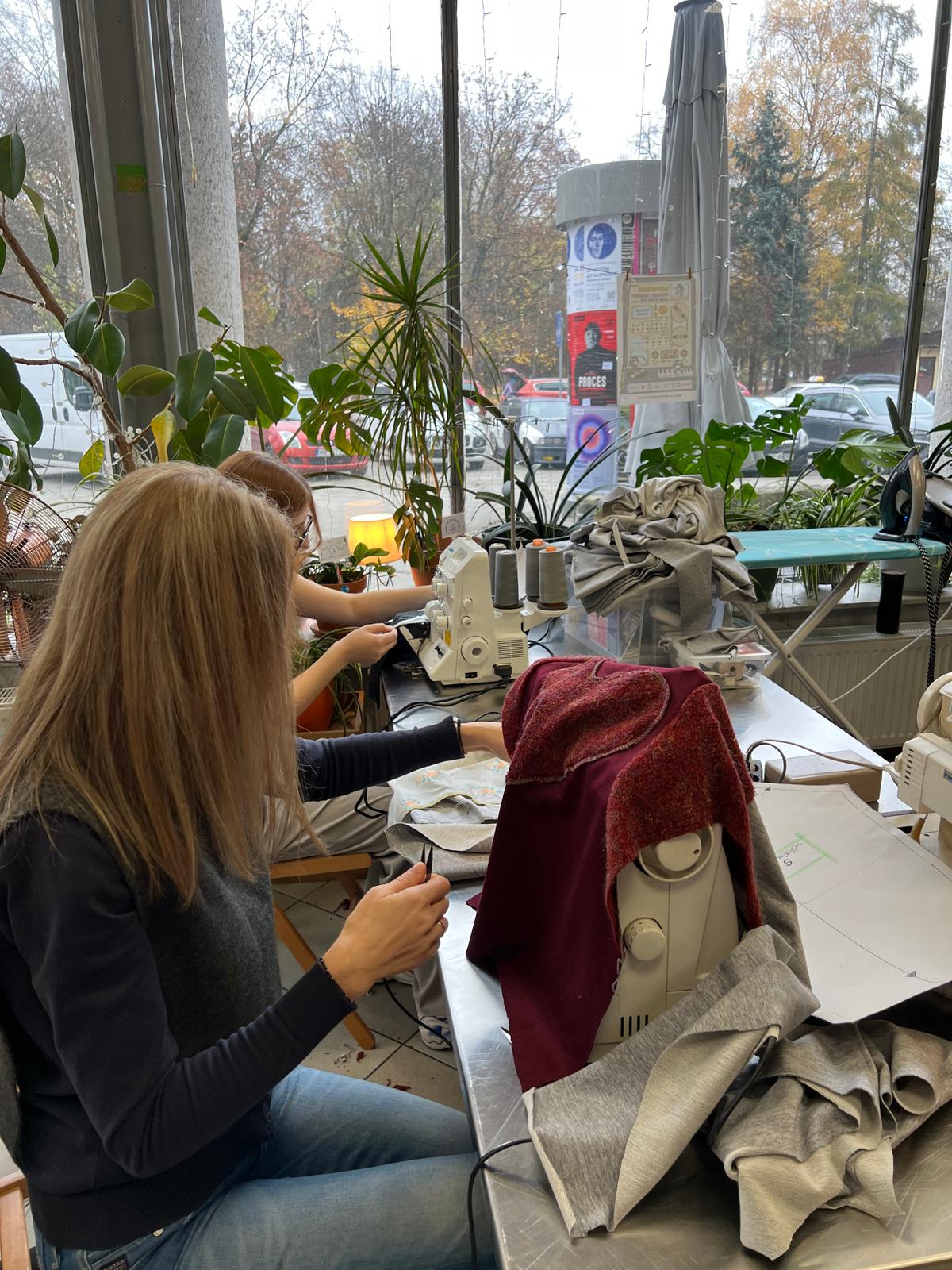 Dziewczyna pracuje przy szyciu na maszynie, siedząc przy oknie z widokiem na jesienną ulicę i rośliny doniczkowe.