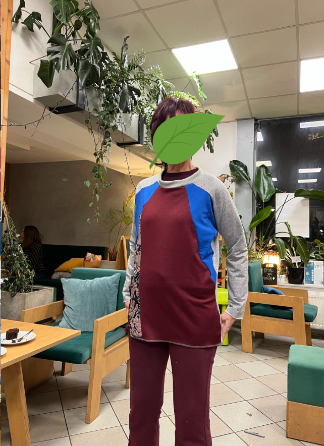 Osoba w kolorowej bluzie stoi w kawiarni otoczonej roślinami doniczkowymi, z dużym zielonym liściem zasłaniającym jej twarz.