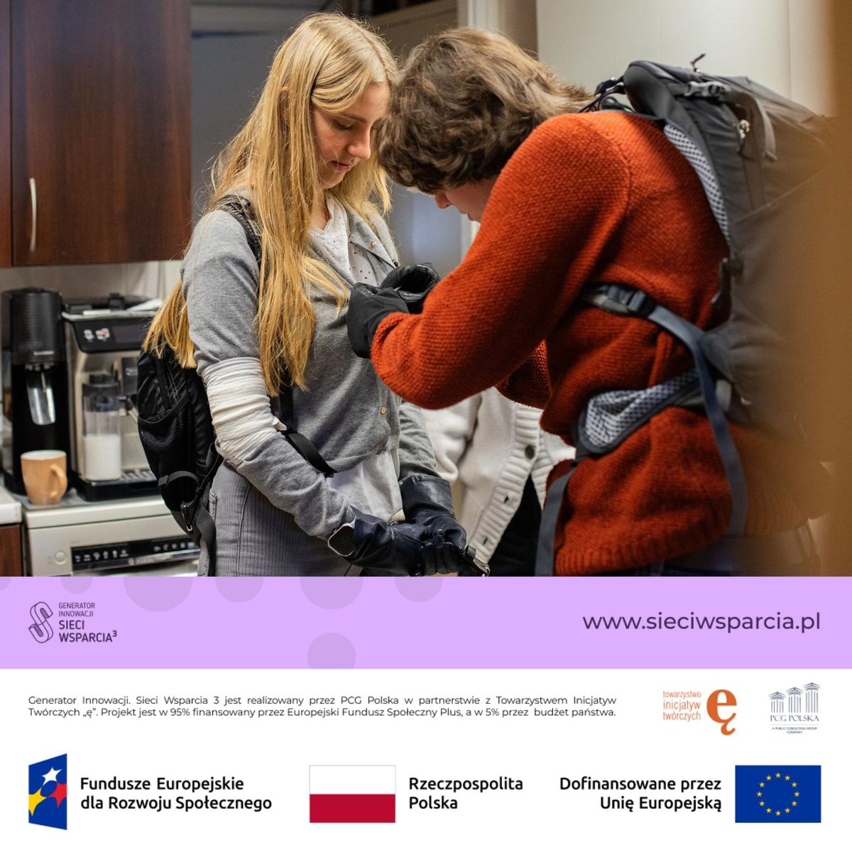 Dwie kobiety rozmawiają w kuchni, jedna ubrana w szarą bluzkę, druga w pomarańczowy sweter, w tle widoczny ekspres do kawy.