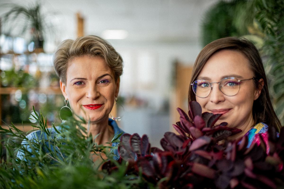 Dwie uśmiechnięte kobiety pozują wśród roślin doniczkowych w jasnym, nowoczesnym wnętrzu.