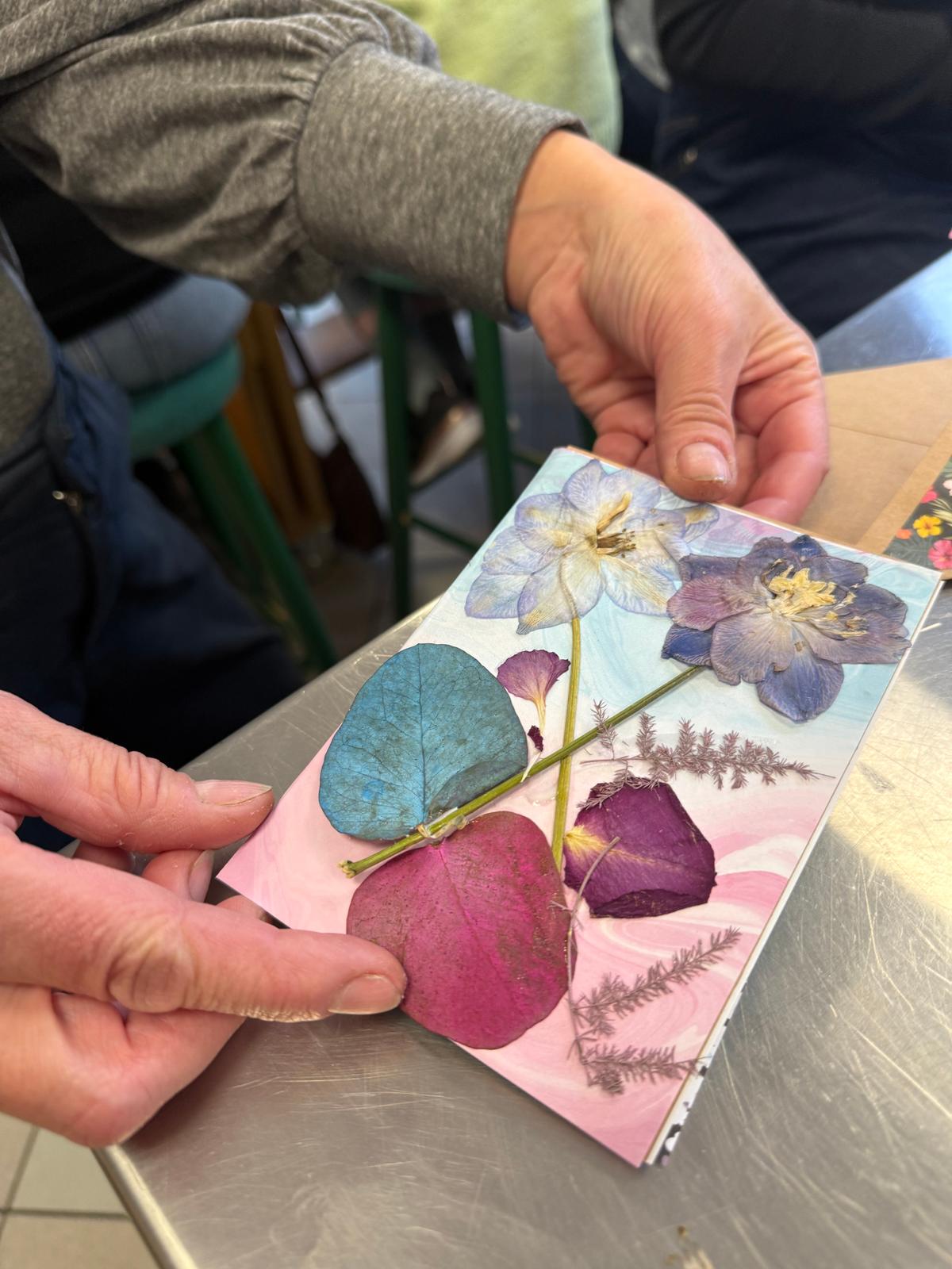 Osoby trzymają kartkę z przetłoczonymi kwiatami i liśćmi w różnych kolorach, pokazując piękne prasowane kwiaty.