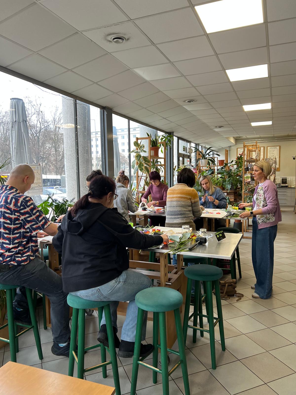 Grupa osób uczestniczy w warsztatach florystycznych w jasnym, słonecznym wnętrzu z roślinami i dużymi oknami.