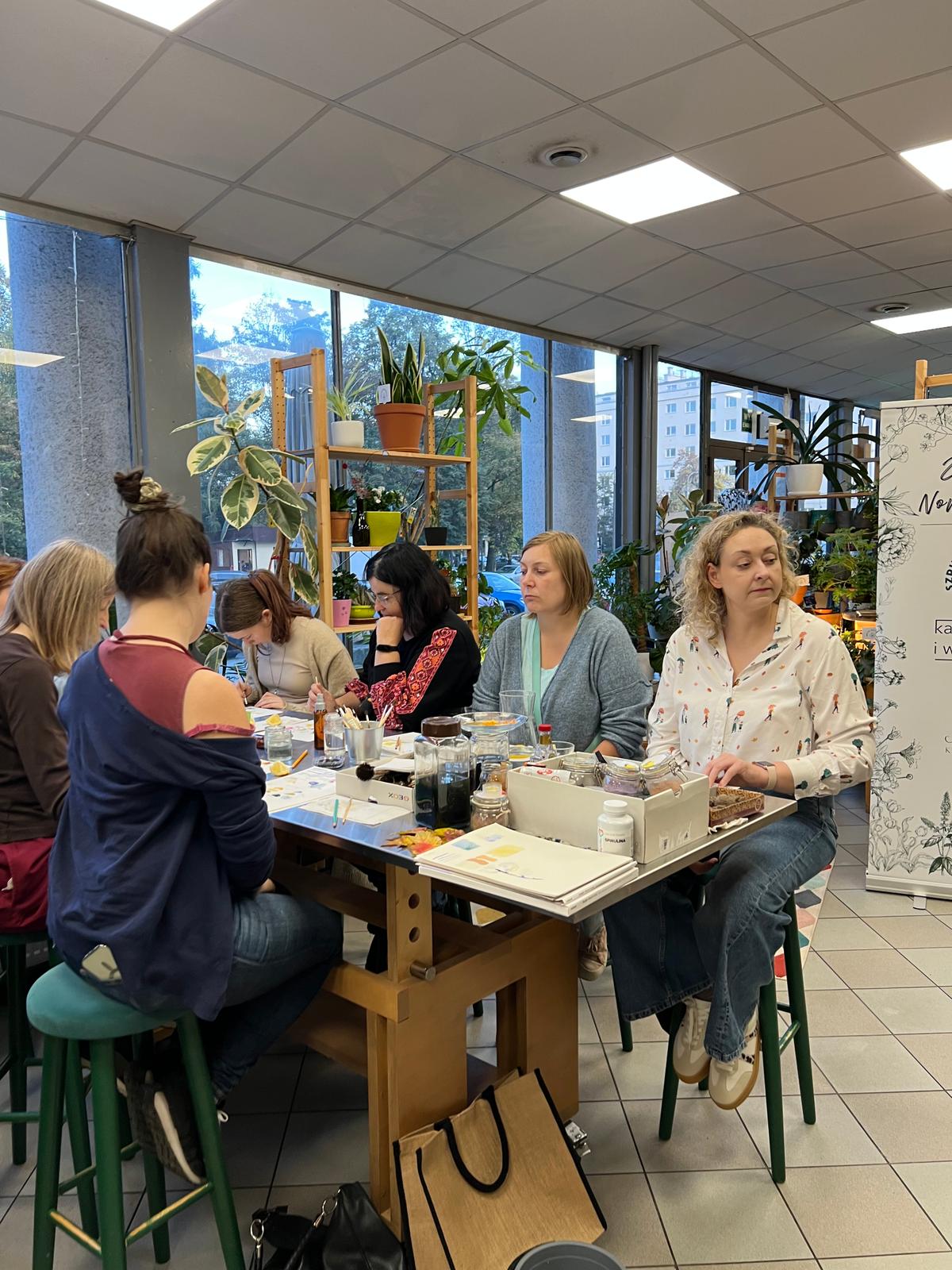 Grupa kobiet uczestniczy w warsztacie lub spotkaniu w jasnym, zazielenionym wnętrzu z dużymi oknami.