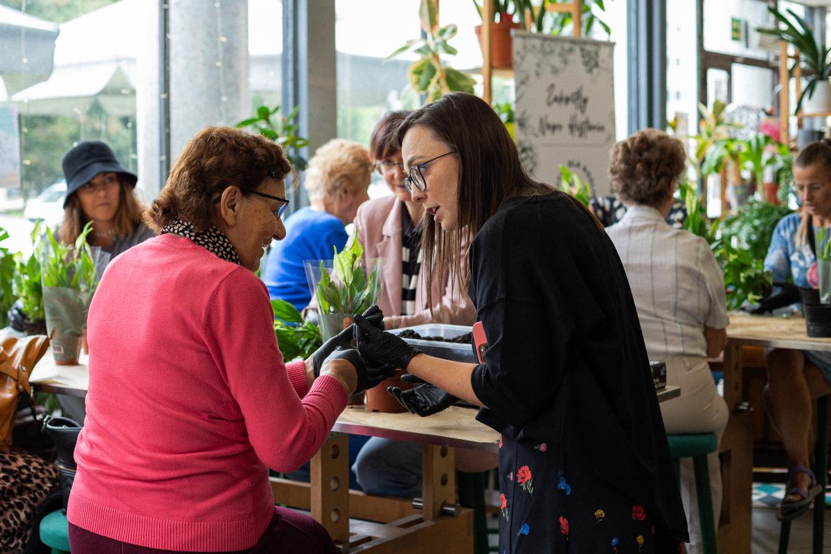 Kobieta w czarnej bluzie rozmawia z uczestniczką warsztatu w otoczeniu roślin doniczkowych.