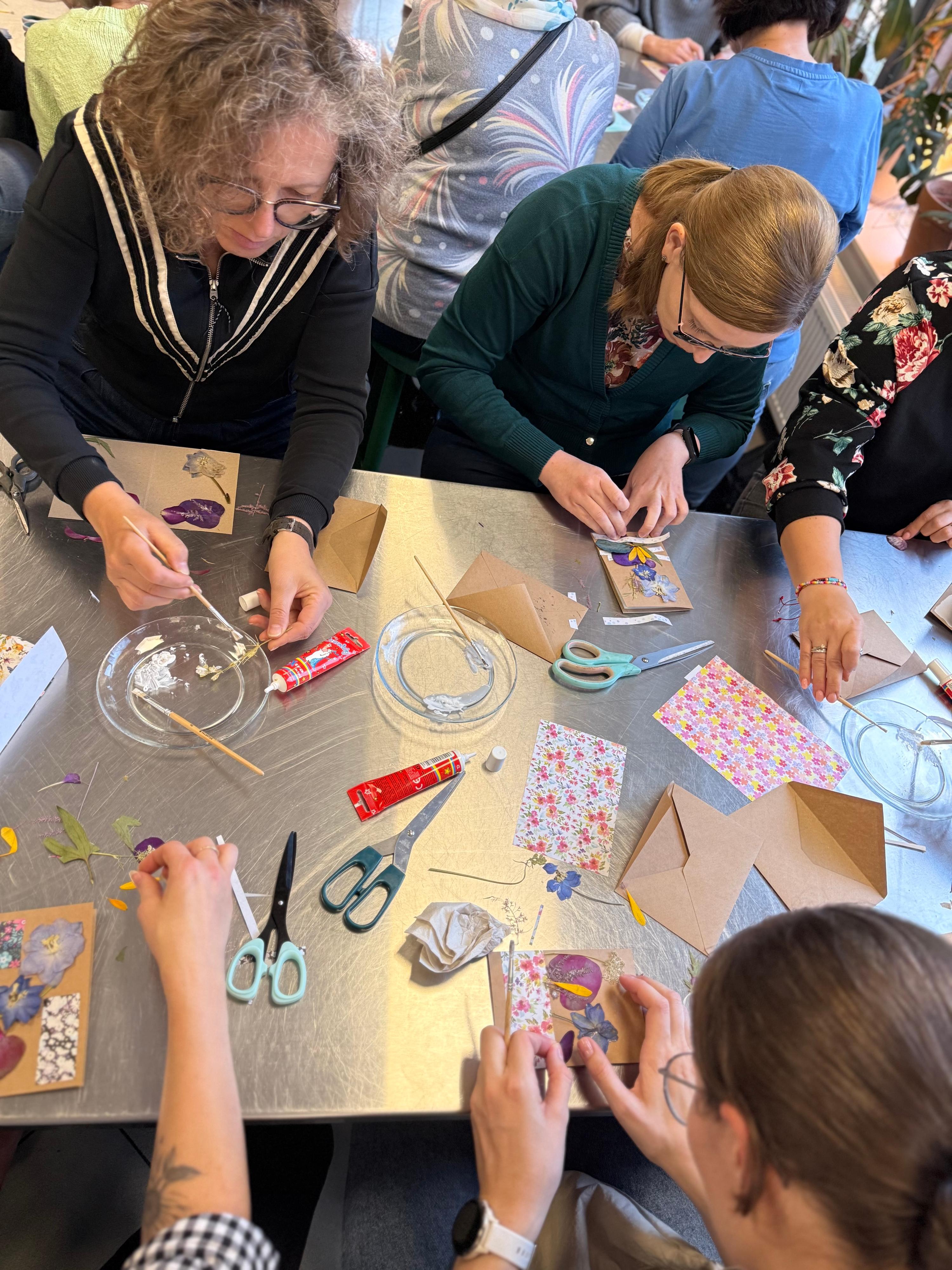 Grupa osób uczestniczy w warsztacie twórczym przy stole, tworząc prace z papieru i materiałów rzemieślniczych.