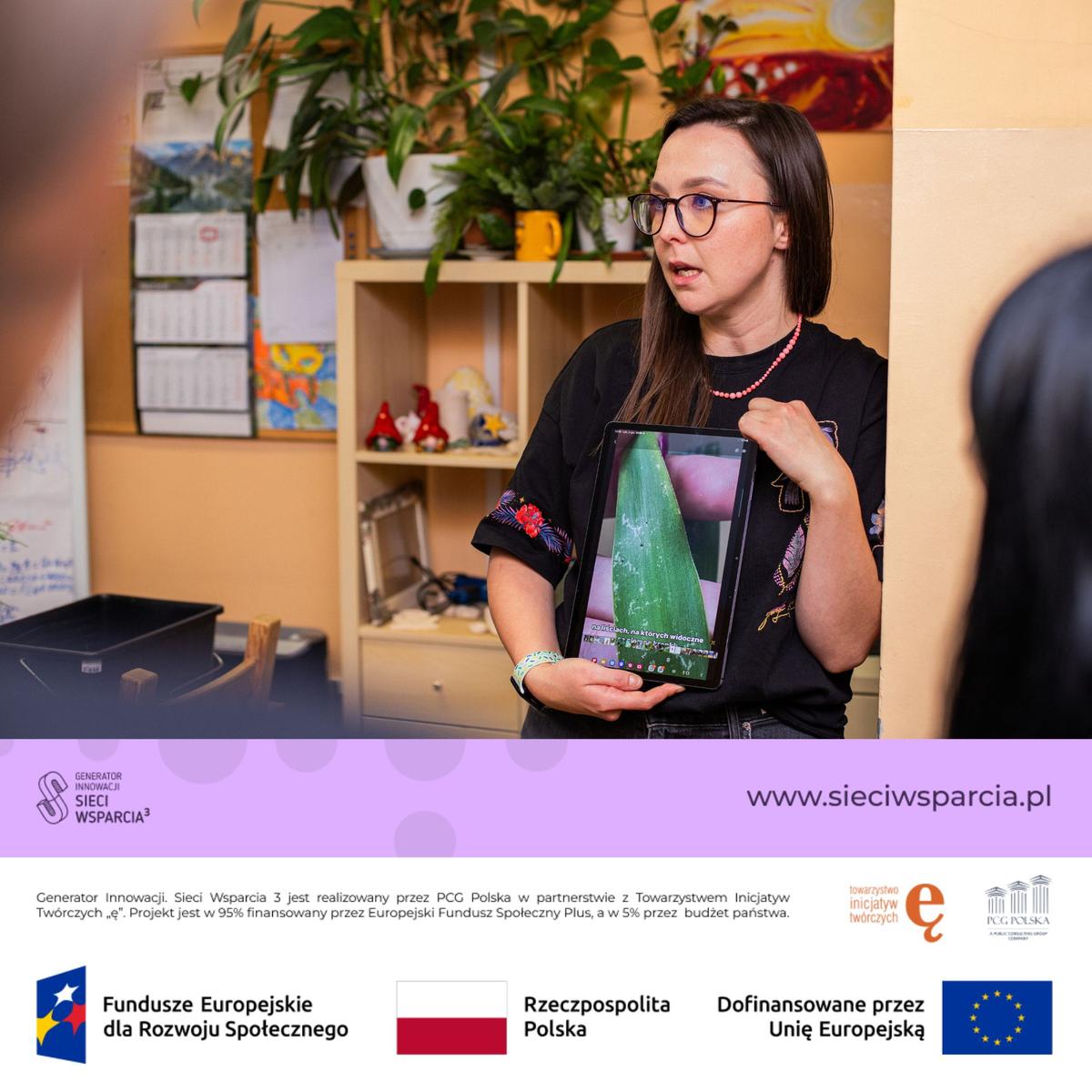 Kobieta w czarnej koszulce i okularach pokazuje tablet podczas prezentacji w pokoju z roślinami.