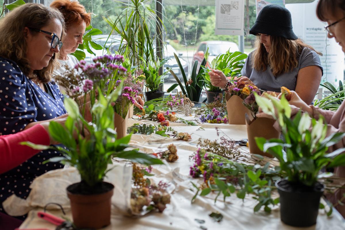Grupa kobiet podczas warsztatu florystycznego przy stole z kwiatami i roślinami w słonecznym pomieszczeniu.