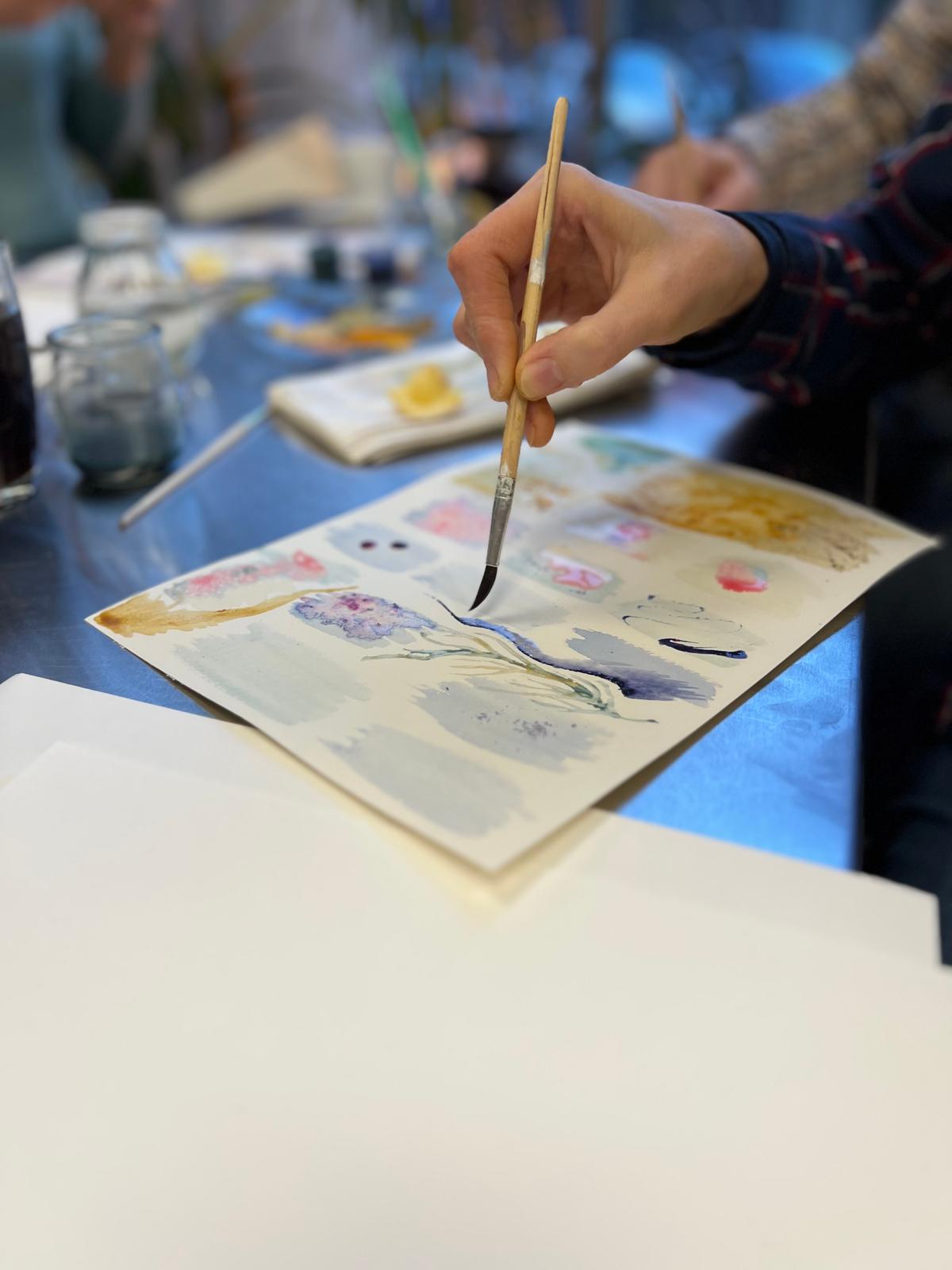 Osoba maluje pędzlem na papierze podczas zajęć artystycznych przy stole z błękitnym obrusem.