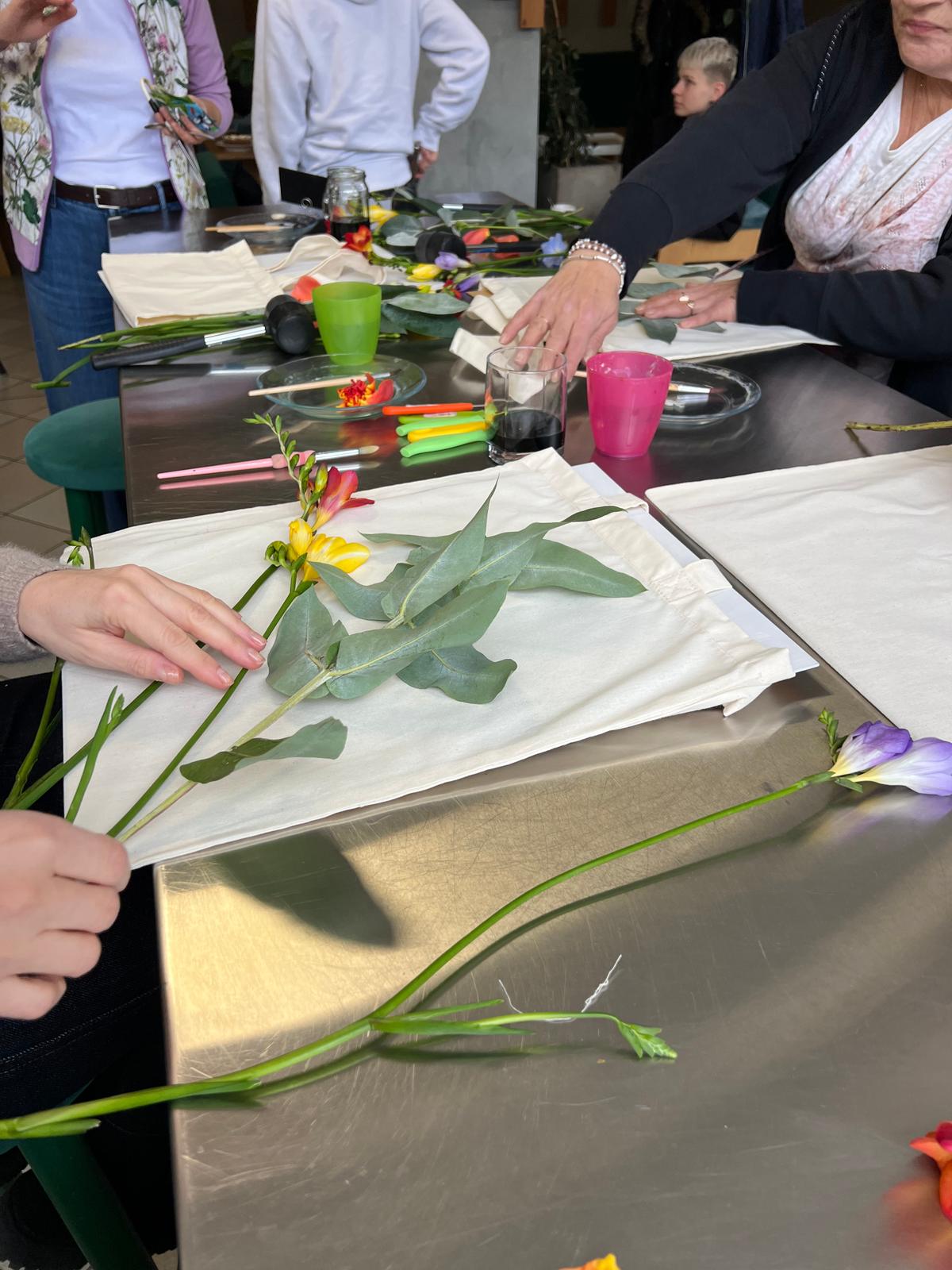 Grupa osób uczestniczy w warsztacie florystycznym, tworząc kompozycje z kolorowych kwiatów i zieleni na białym papierze.