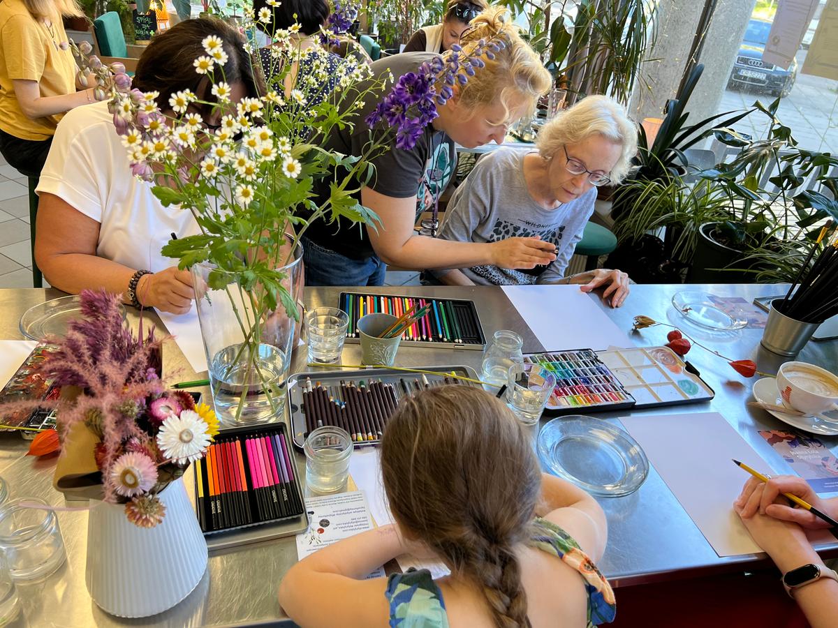 Grupa osób uczestniczy w warsztatach artystycznych przy stole z kwiatami, kredkami i papierem do rysowania.