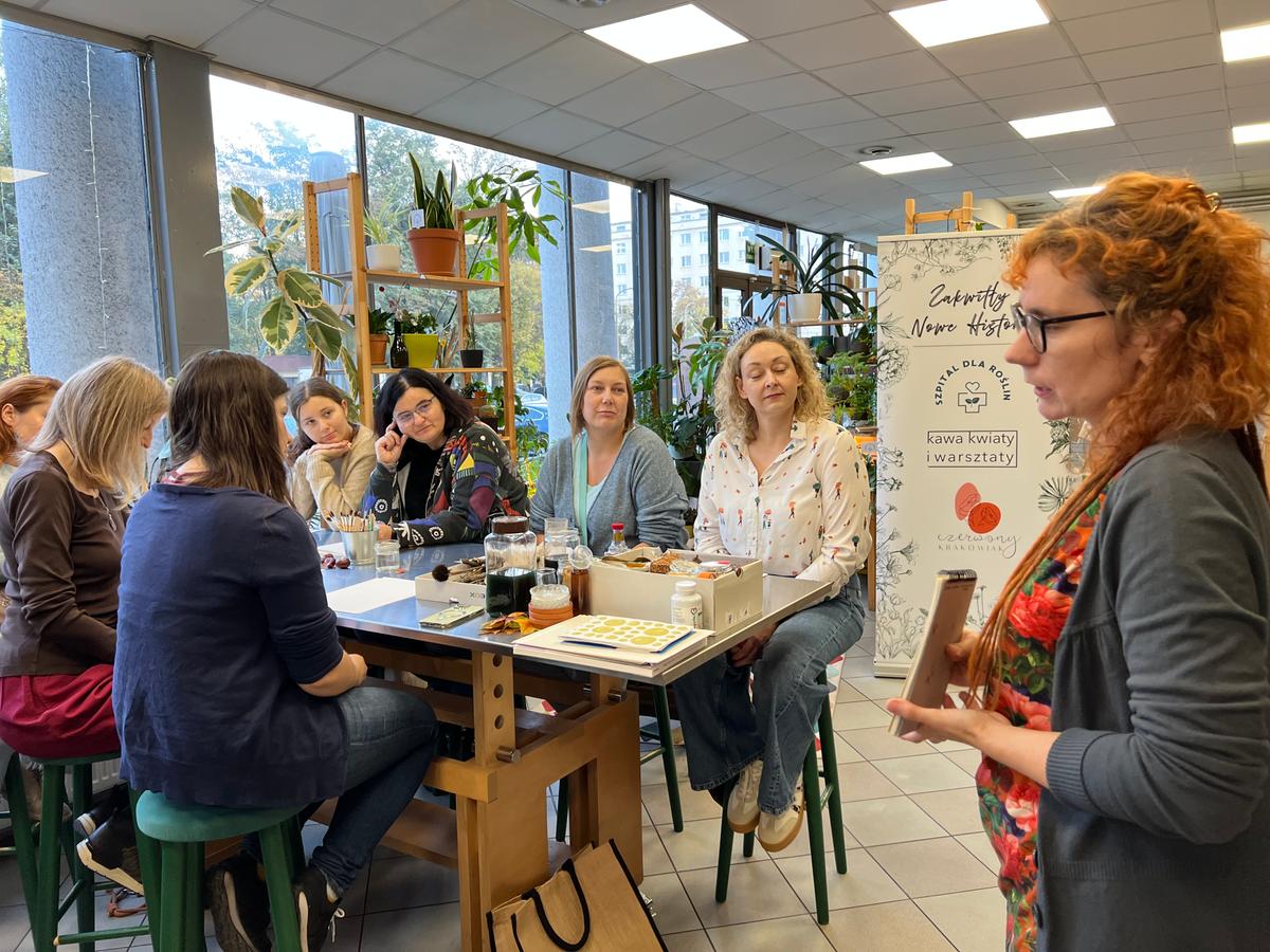 Grupa kobiet uczestniczy w warsztatach lub spotkaniu edukacyjnym w nowoczesnym pomieszczeniu z roślinami i dużymi oknami.