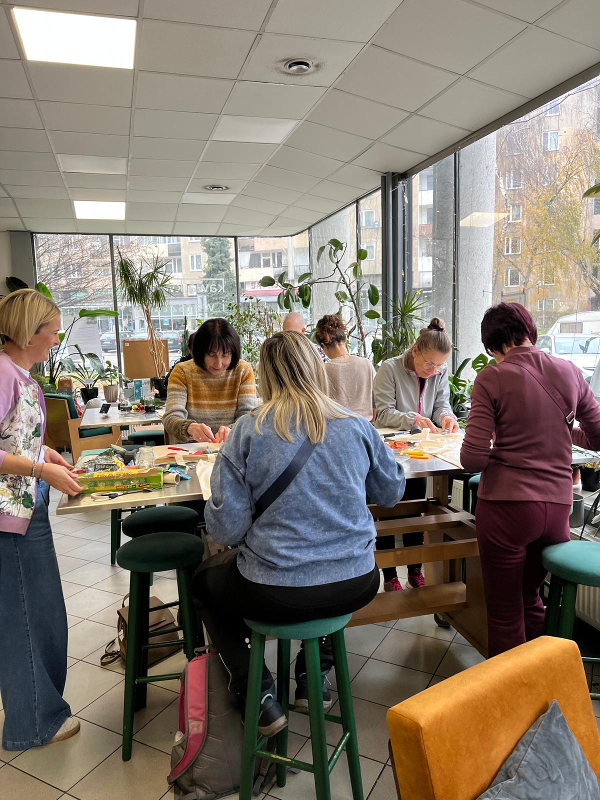 Grupa ludzi uczestniczy w zajęciach artystycznych przy stołach w jasnej sali z dużymi oknami i roślinami.