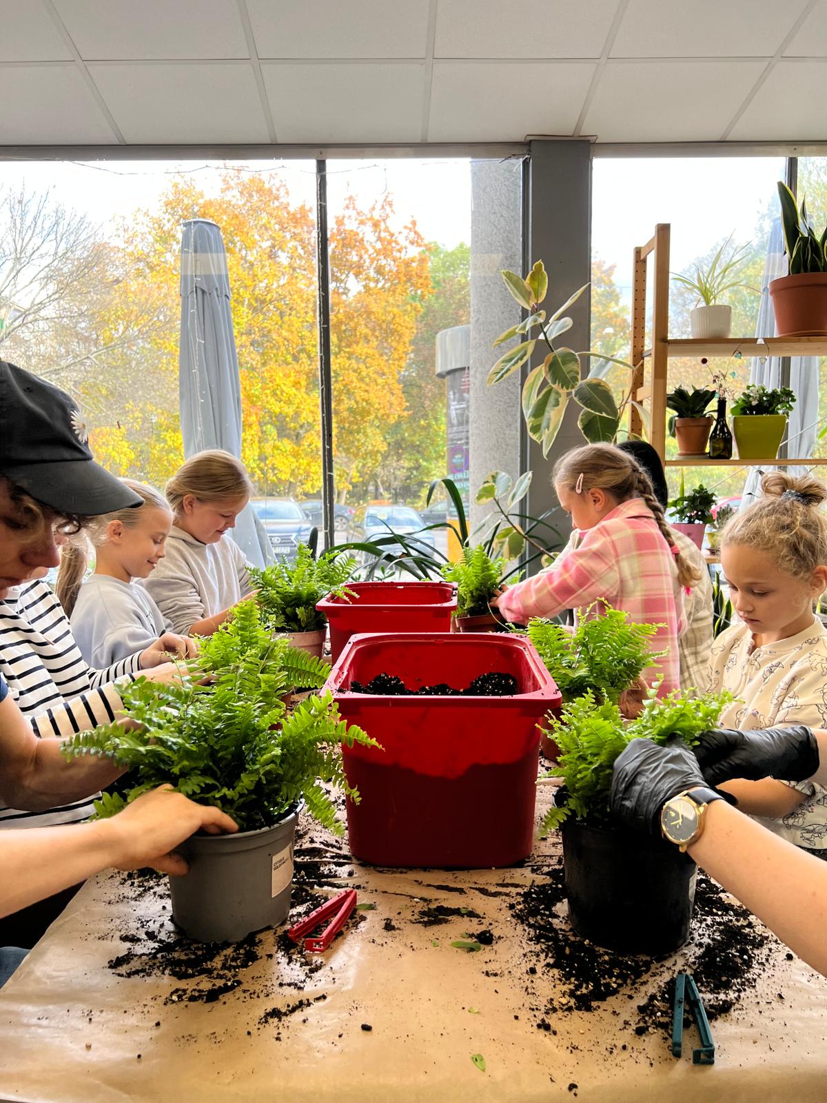 Grupa dzieci pracuje przy sadzeniu roślin w doniczkach, wykonując zajęcia ogrodnicze w jasnym pomieszczeniu.