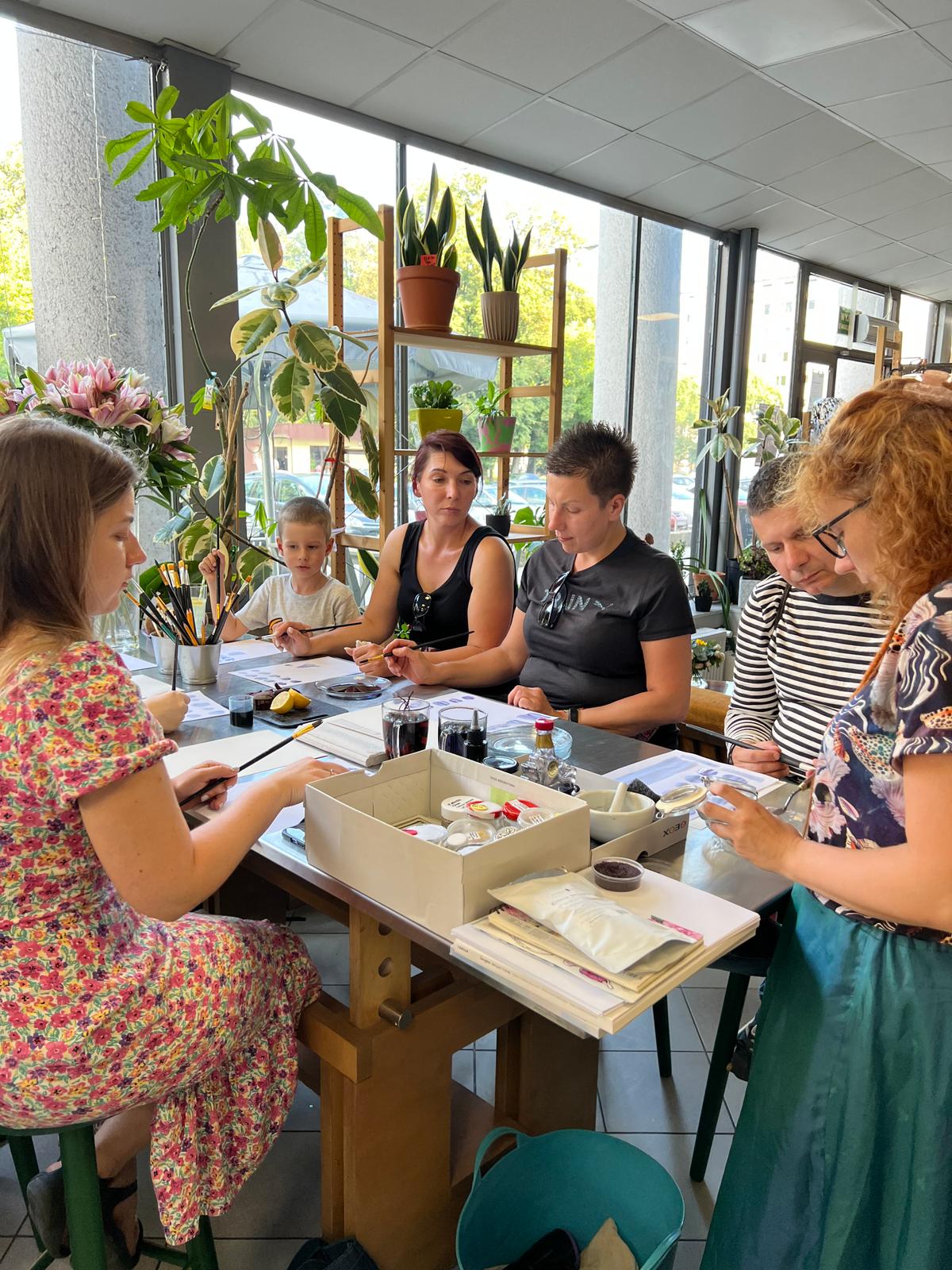 Grupa osób uczestniczy w warsztacie plastycznym w jasnej sali pełnej roślin przy dużych oknach.