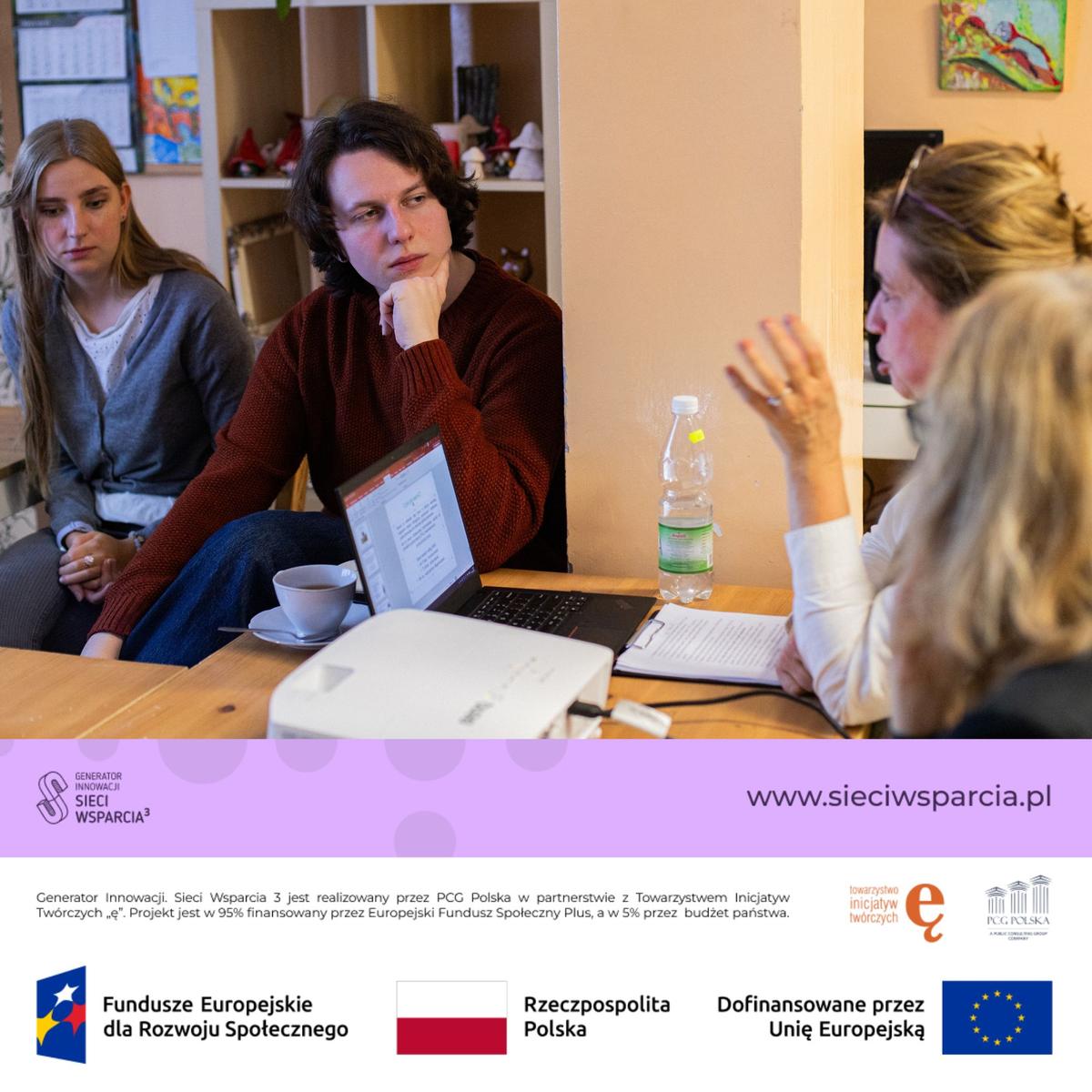 Trzy kobiety uczestniczą w spotkaniu biznesowym przy stole z laptopem i materiałami roboczymi.