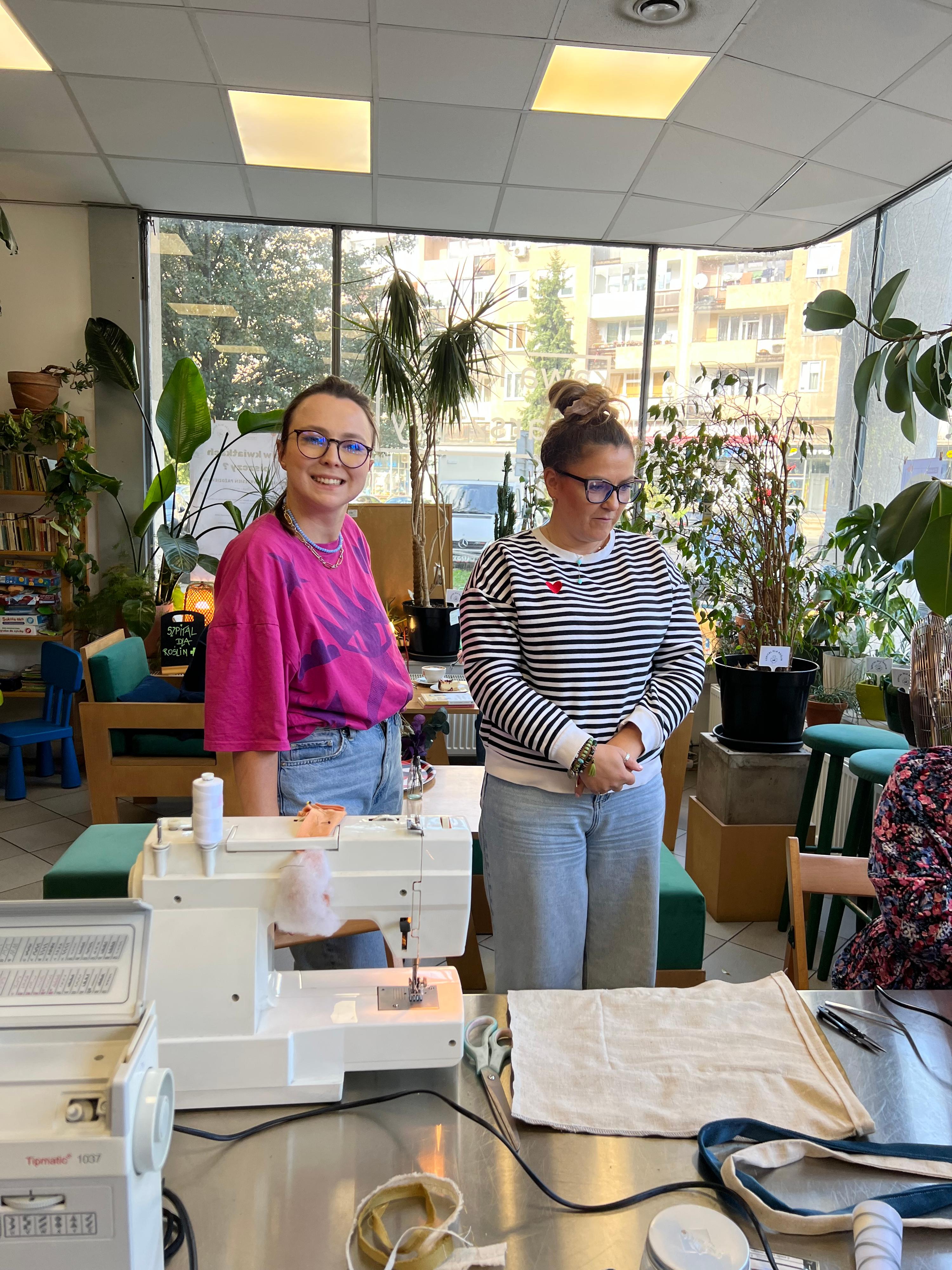 Dwie dziewczyny w pracowni krawiecko-projektowej otoczone roślinami i maszynami do szycia.