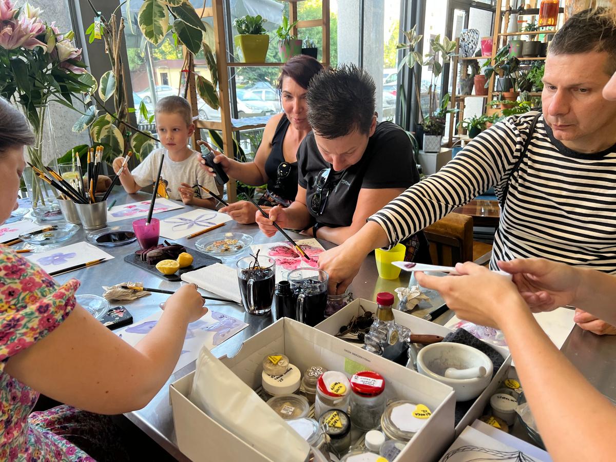 Grupa ludzi uczestniczy w warsztacie malarskim w otoczeniu roślin, malując akwarelami przy stole z art supplies.