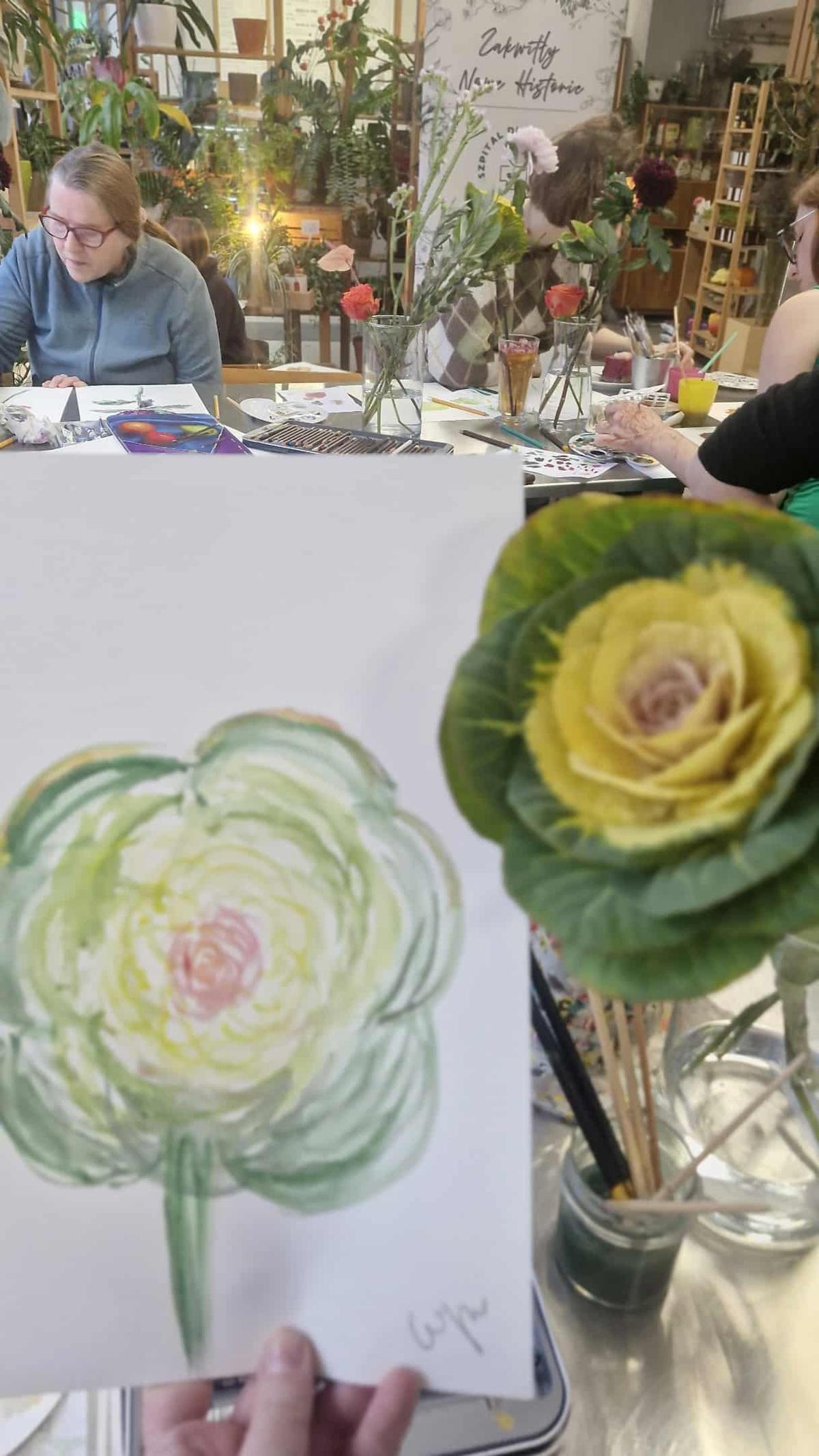 Grupa osób uczestniczy w warsztacie malarskim w pięknie urządzonym studio pełnym roślin, rysując kwiaty.