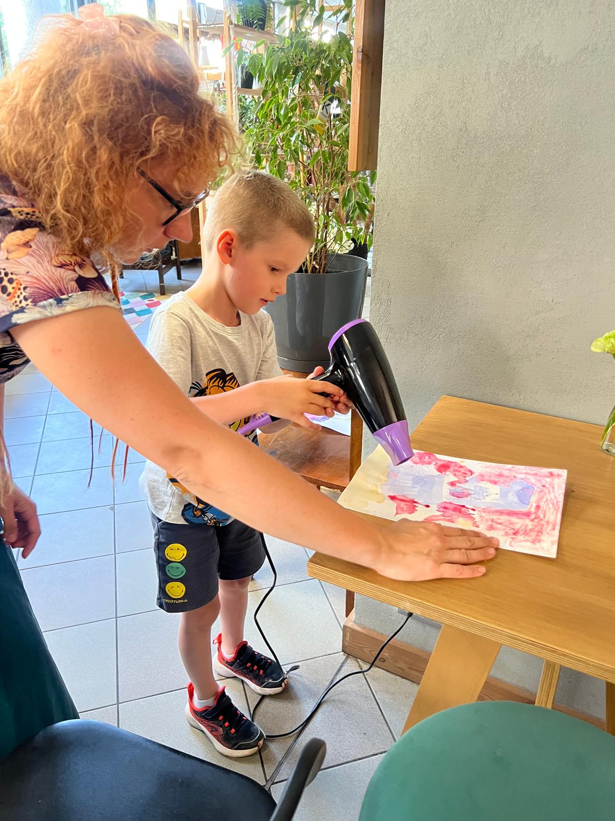 Dorosła kobieta pokazuje chłopcu na tablecie obrazek z kwiatami, siedząc przy drewnianym stole w jasnym pomieszczeniu.