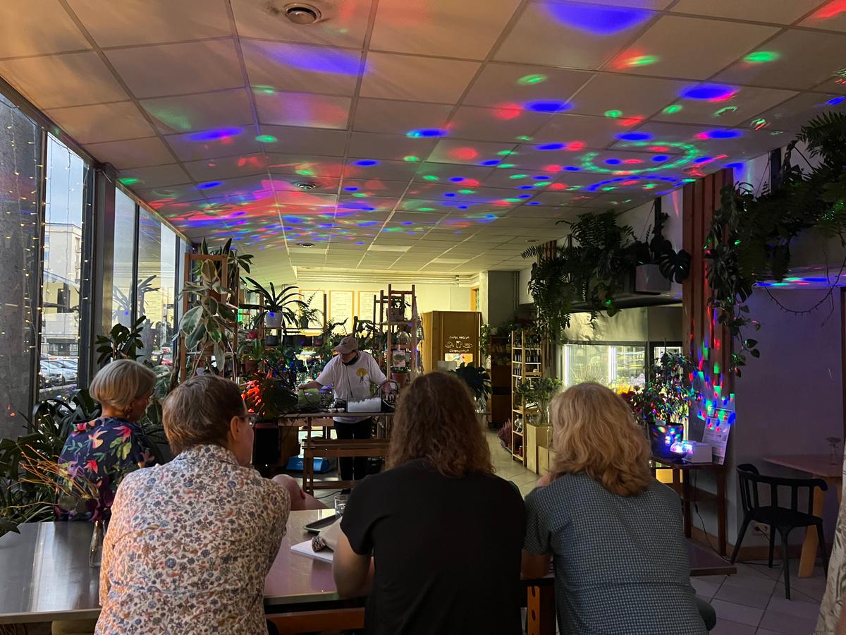 Trzy osoby sieddzą w kwiaciarni z kolorowymi światłami na suficie, słuchając mężczyzny stojącego za ladą.