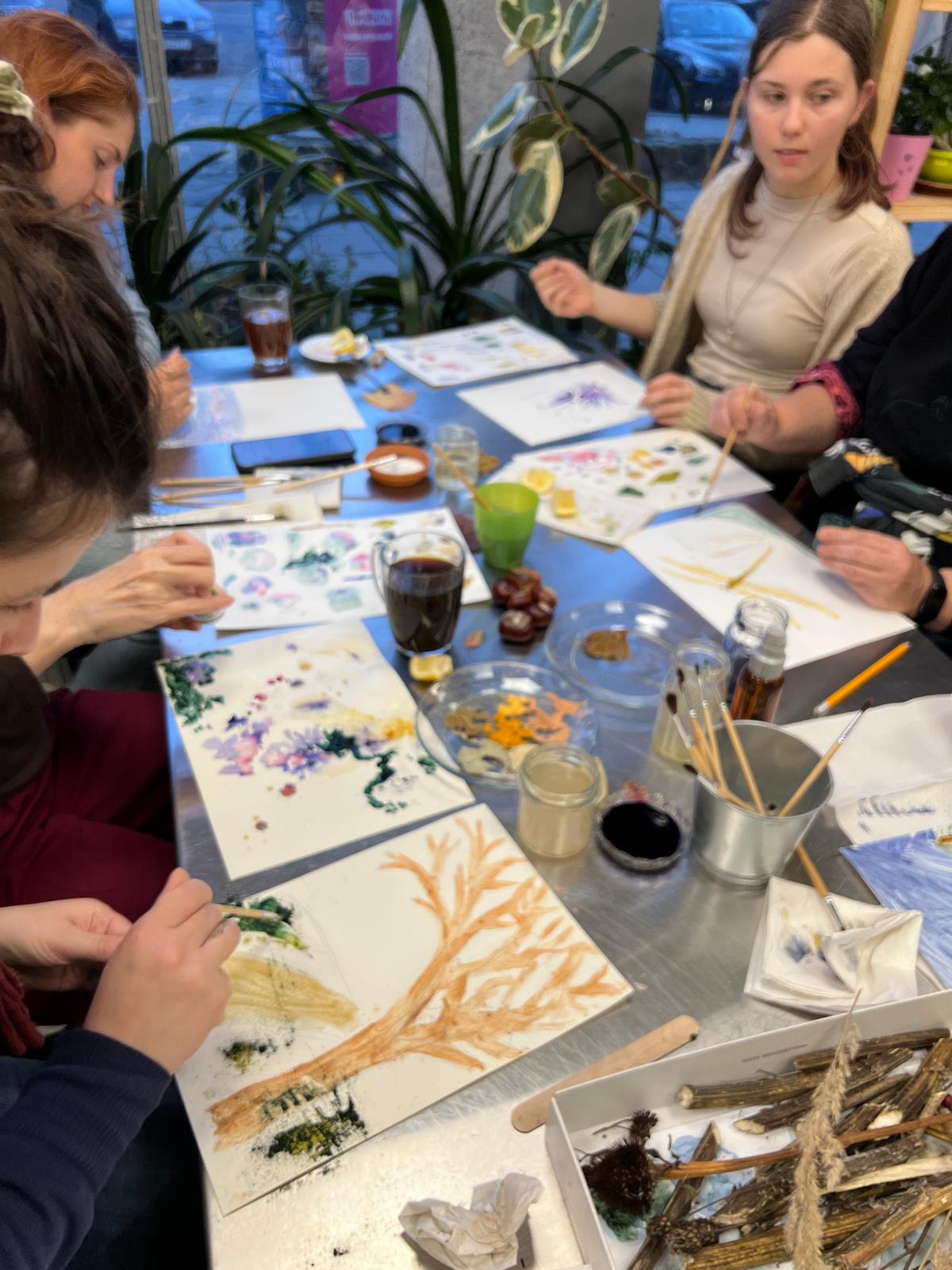 Osoby uczestniczą w warsztacie malarskim, tworząc akwarelowe prace inspirowane naturą przy stole pełnym materiałów artystycznych.