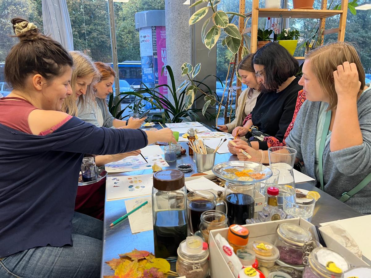 Grupa dziewczyn uczestniczy w warsztatach artystycznych przy stole z materiałami do malowania w jasnym pokoju z roślinami.