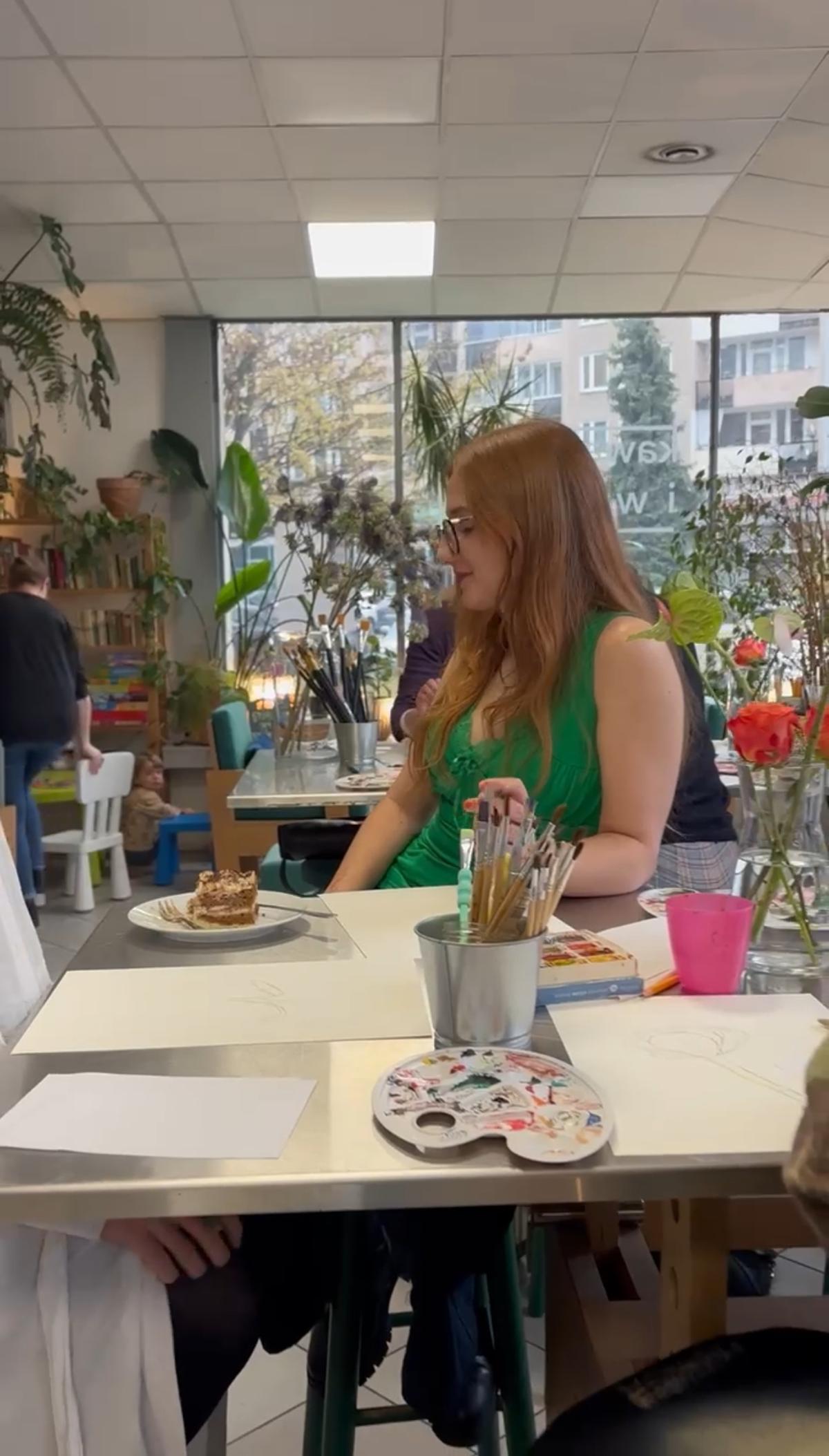 Kobieta w zielonej sukience siedzi przy stole w jasnym studio z roślinami, jedzeniem i artystycznymi materiałami.